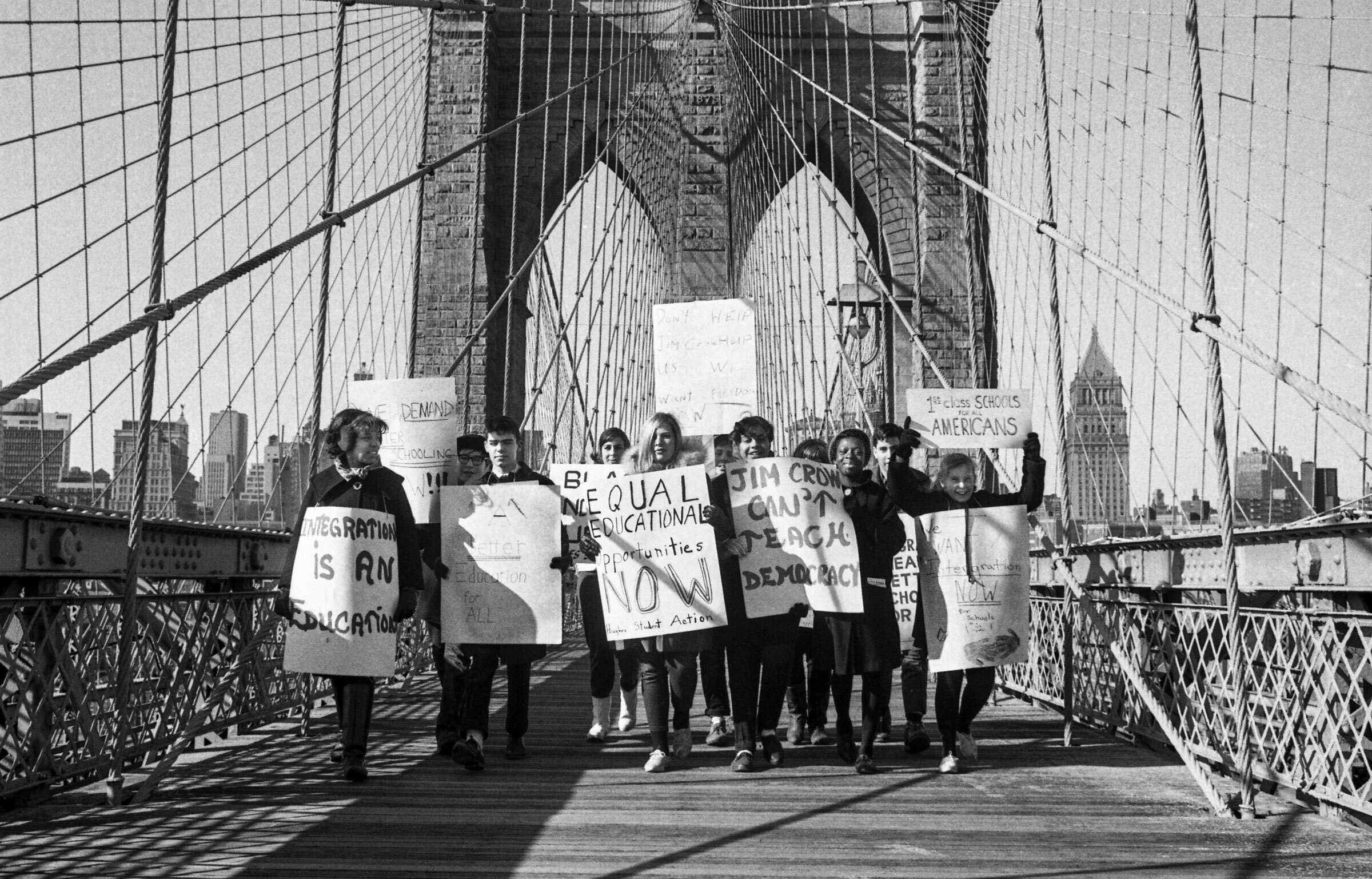 Black and white photograph of students and teachers standing on a bridge with protest signs decrying school segregation.