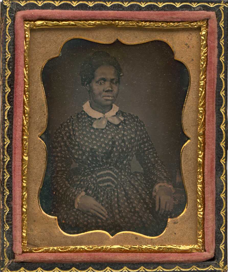 A 1/9th plate daguerreotype portrait of Rhoda Phillips (1831-1906), a woman who was born enslaved and owned by the Clark-Gleaves family of Nashville, Tennessee, with whom she remained after emancipation. Phillips is seated in the image and wears a dress with a flower pattern, lace cuffs, and a lace collar. The case is contemporary and has a decorative gold metal frame and a red velvet lining.