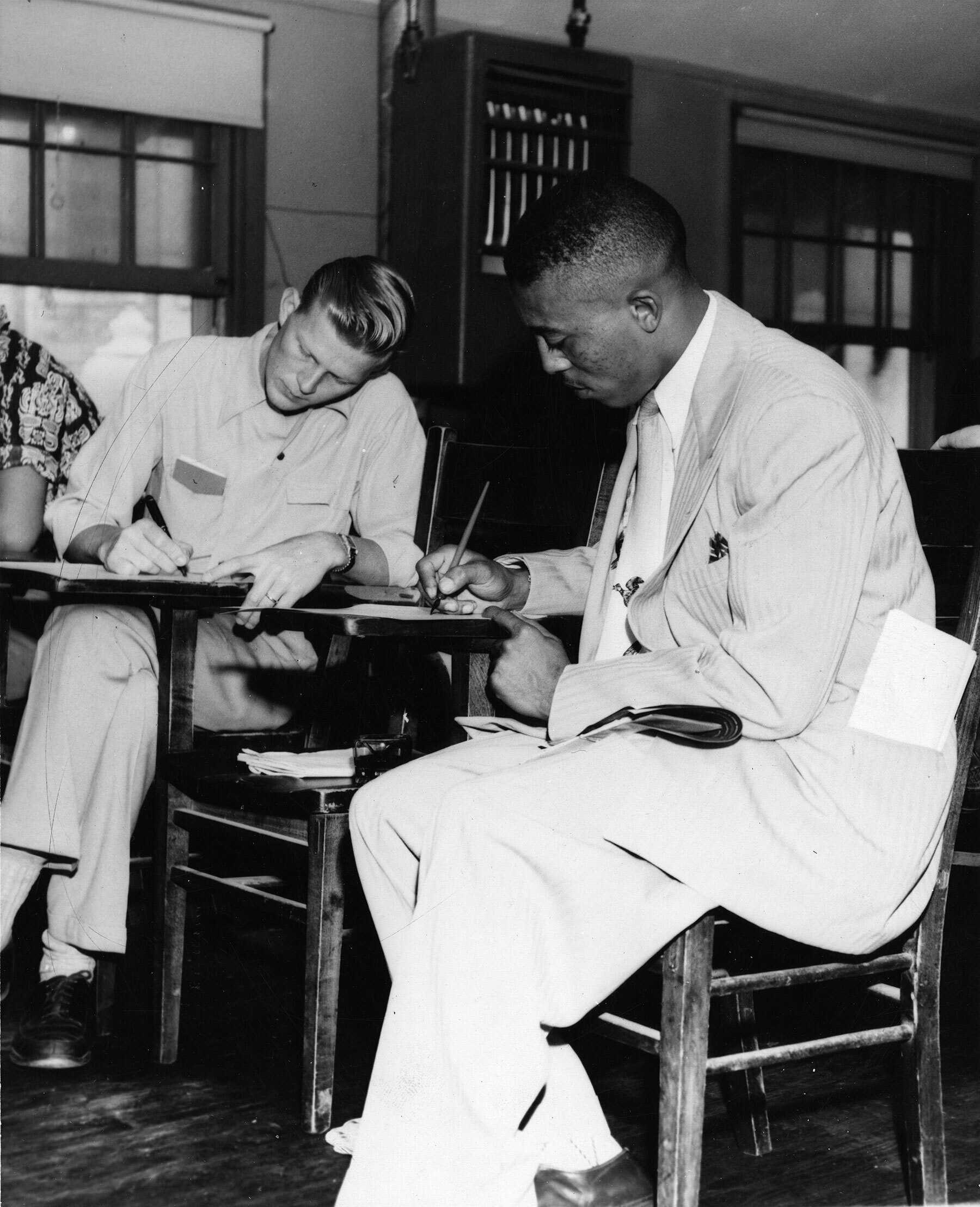 Two people including John Chase (right) writing at desks in an indoor setting.
