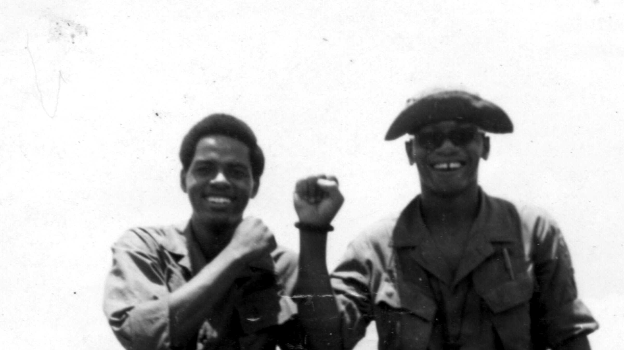 A black and white photograph of two soldiers, in military fatigues, perched on the edge of the windshield, boots resting on the jeep's hood.