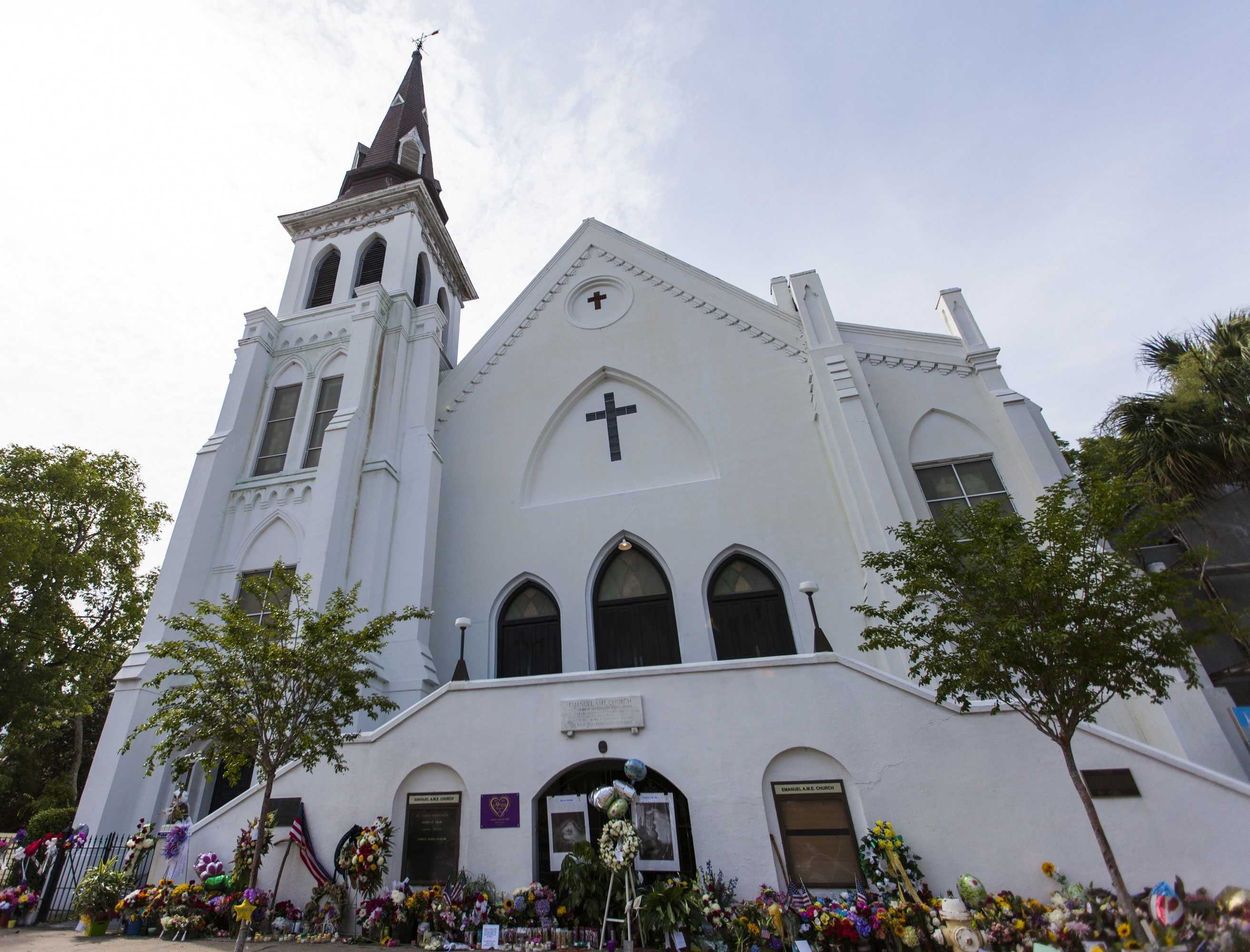 The large white church,Emanuel AME Church, has flowers, pictures, and candles are laid in front.