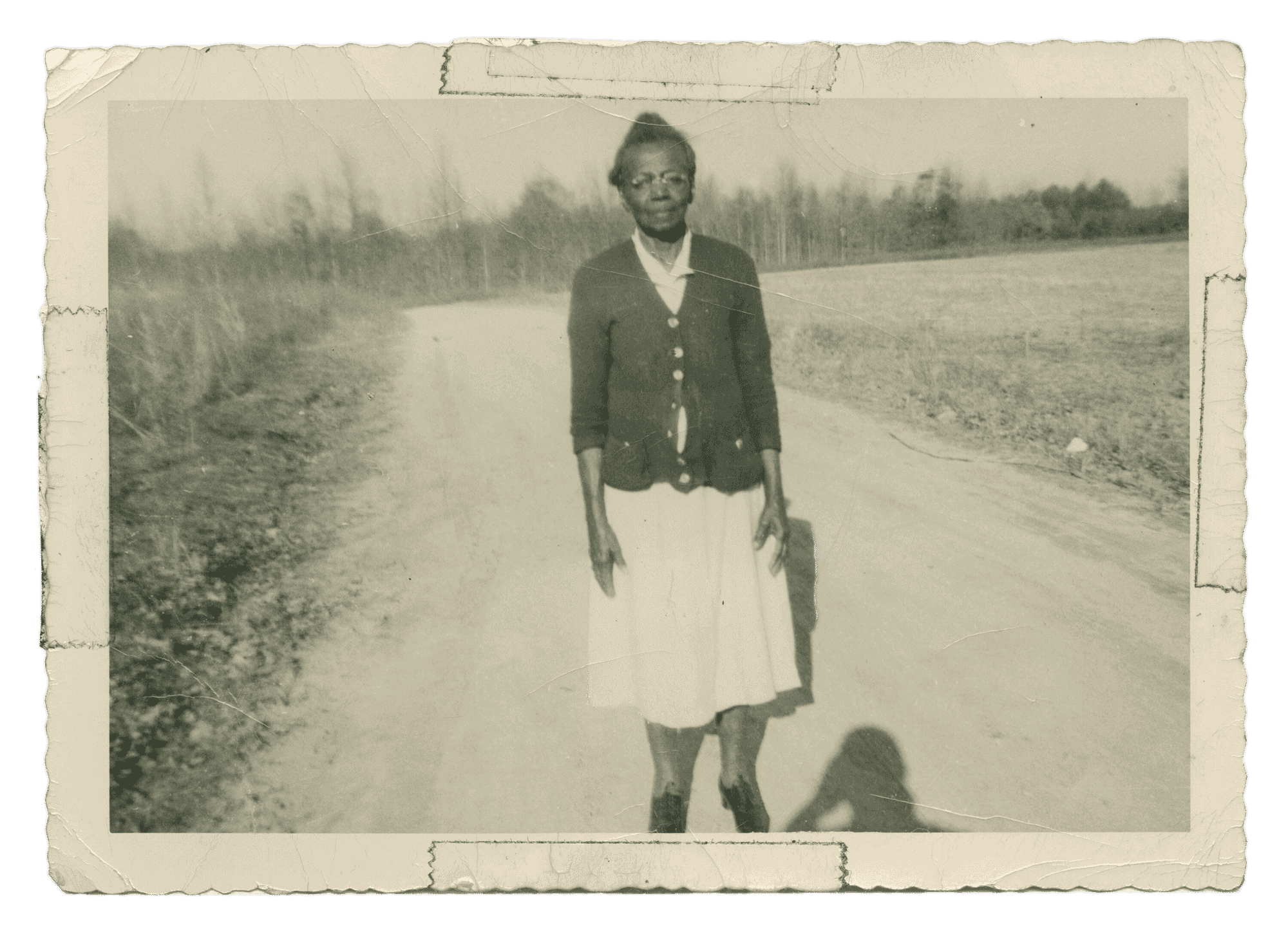 A black-and-white photograph of third-generation midwife Susie Carey, standing on a dirt road in a rural landscape.