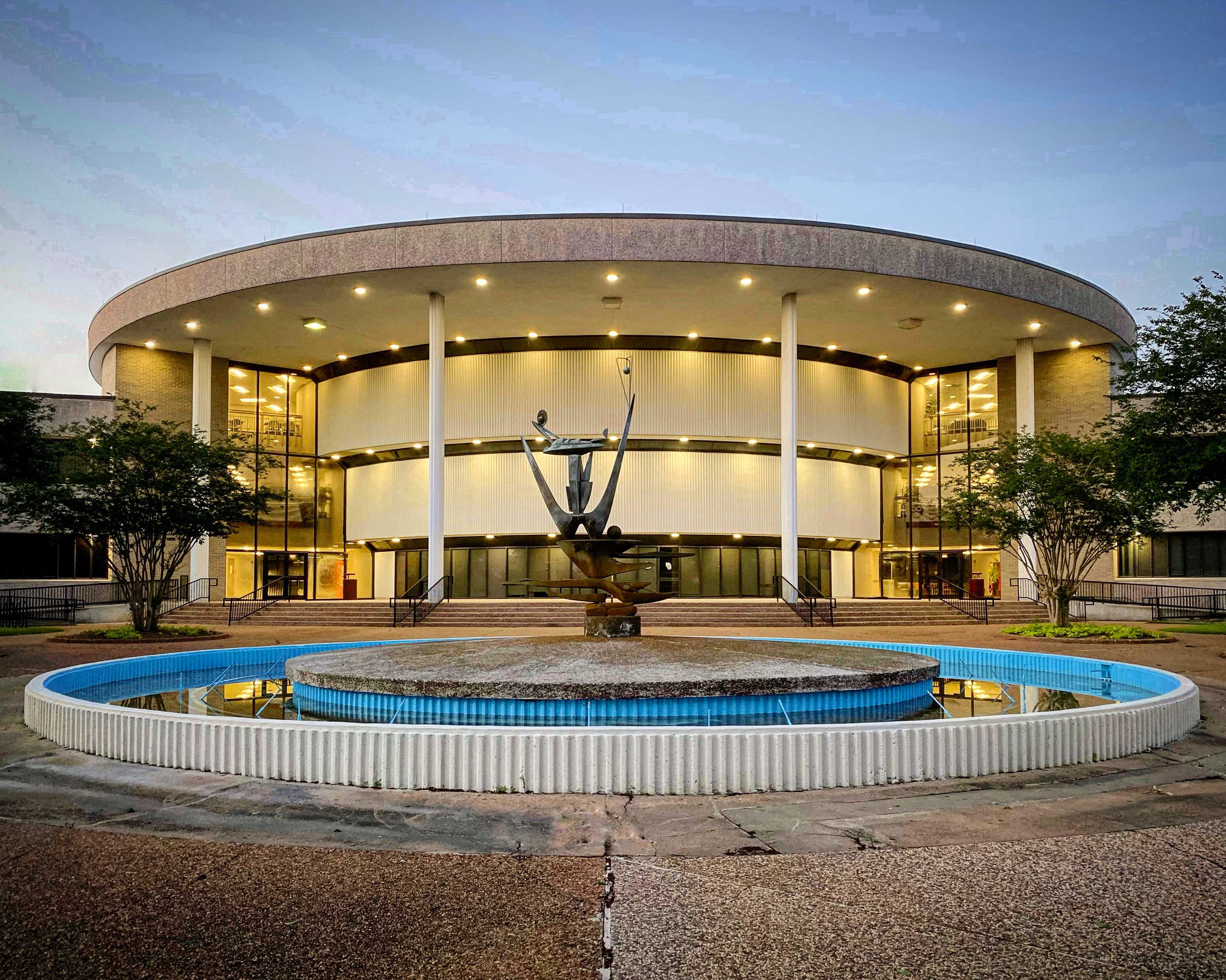 Two-story circular building with large windows and a lit fountain with abstract sculpture in front.