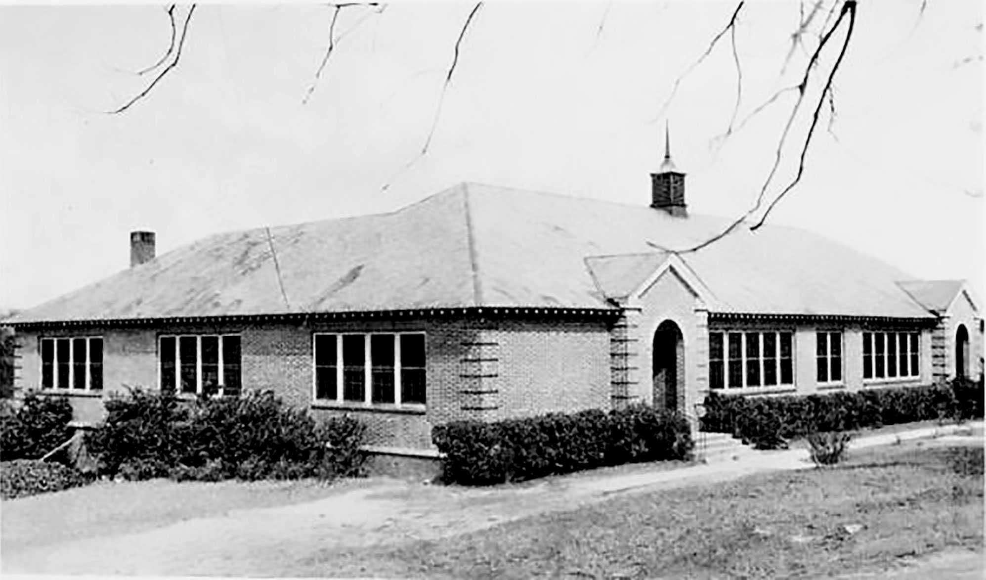 Photo of a single-story brick building with a steep roof and arched entrance, surrounded by bushes.