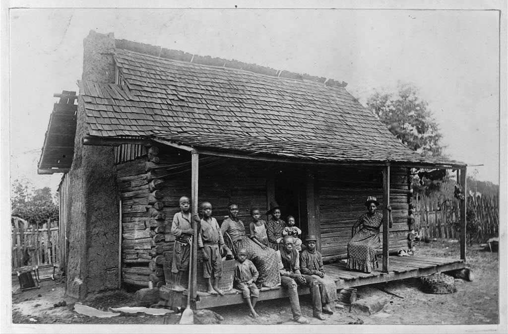A black and white photograph of a group of people sitting on the porch of a cabin-style home.