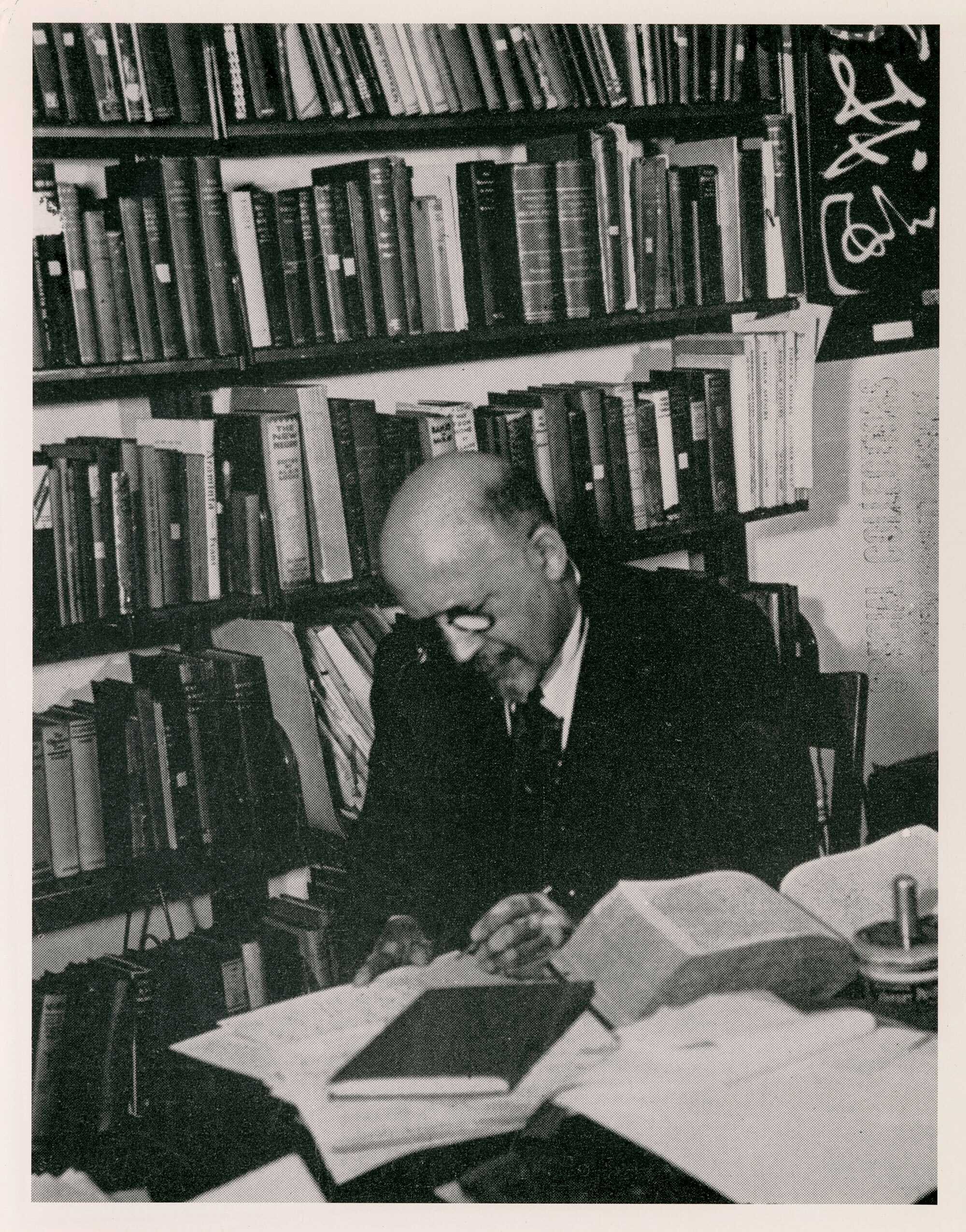 Black and white image of an W.E.B. Du Bois wearing glasses and a suit, seated at a desk cluttered with books and papers, surrounded by bookshelves in a study or office setting