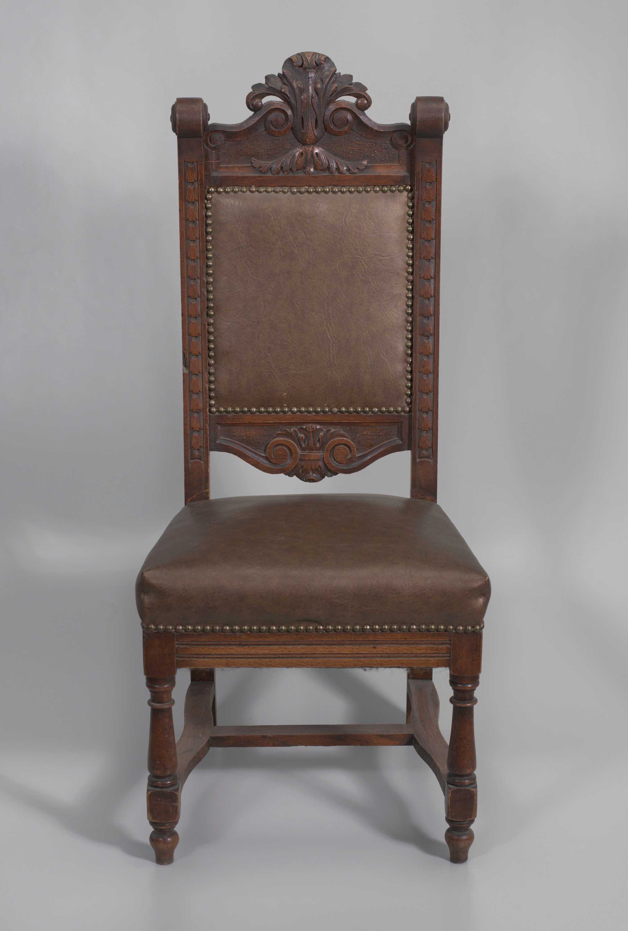 A wood pulpit chair with a dark finish. The chair has four legs with the rear legs extending the full height of the seat back. The front legs are turned with varying geometric shapes and are topped with the seat. Connecting the front legs to the back legs are two curved stretcher bars. Between the two stretcher bars is an additional support bar. The seat cushion and chair back are padded with faux-leather covers. The covers are adhered by metal studs. The chair back features a line of stylized foliage carved on the front of either side of the uprights. The uprights of the chair back are capped with scrolls. The center of the top rail of the chair features large, carved acanthus leaves in the center with scrollwork extending down either side. The bottom rail of the chair back features a similar but smaller design of acanthus leaves and scrollwork.