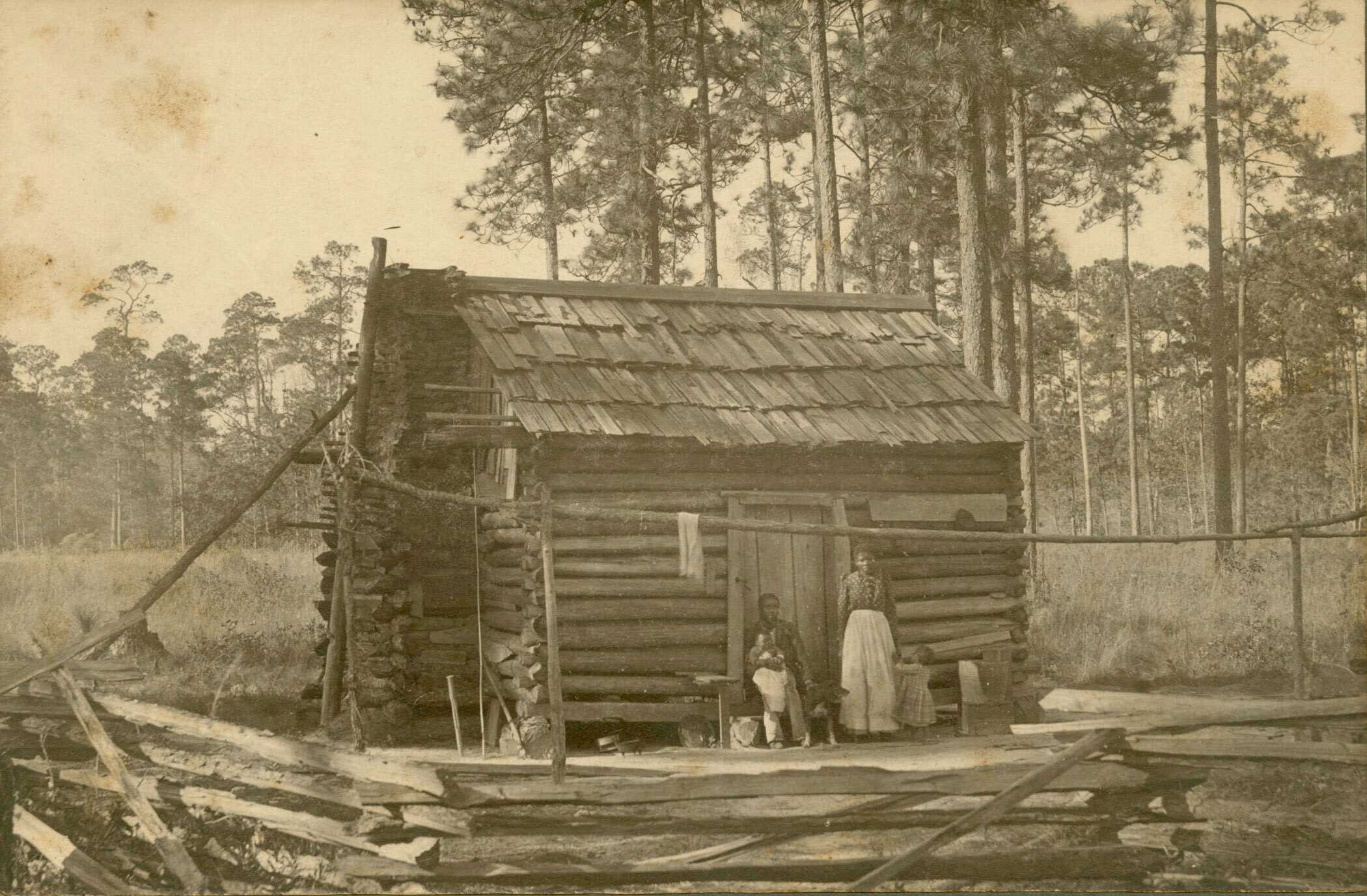 Black and White photograph of a family in front of a log cabin with dog.