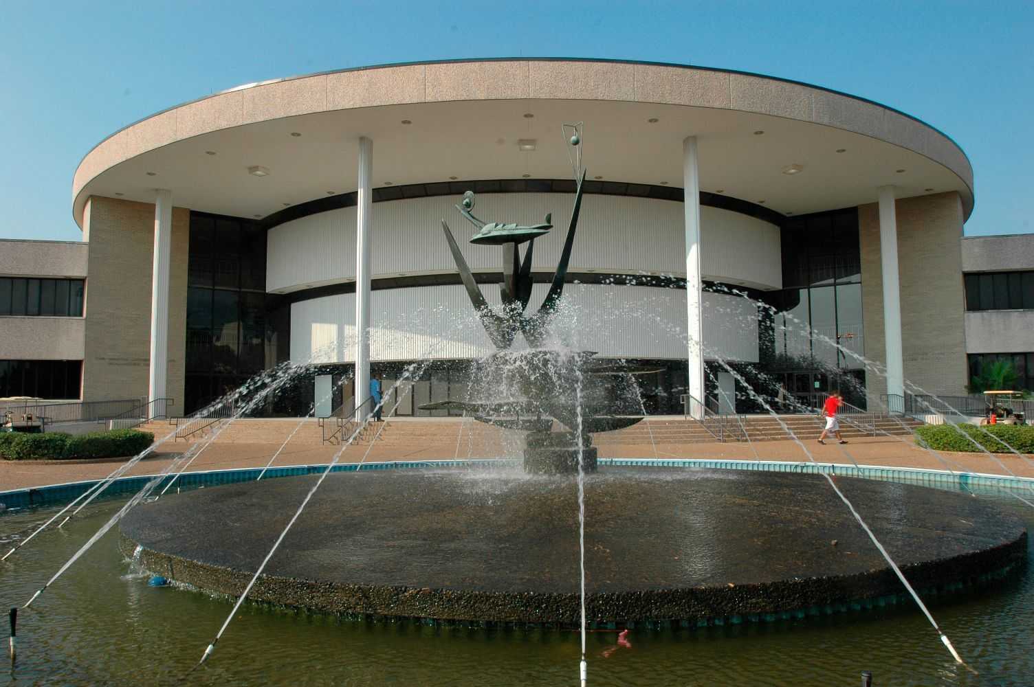 Modern circular building with tall white columns and an overhanging roof, fronted by a large circular fountain with upward-shooting water streams and an abstract metal sculpture at its center.