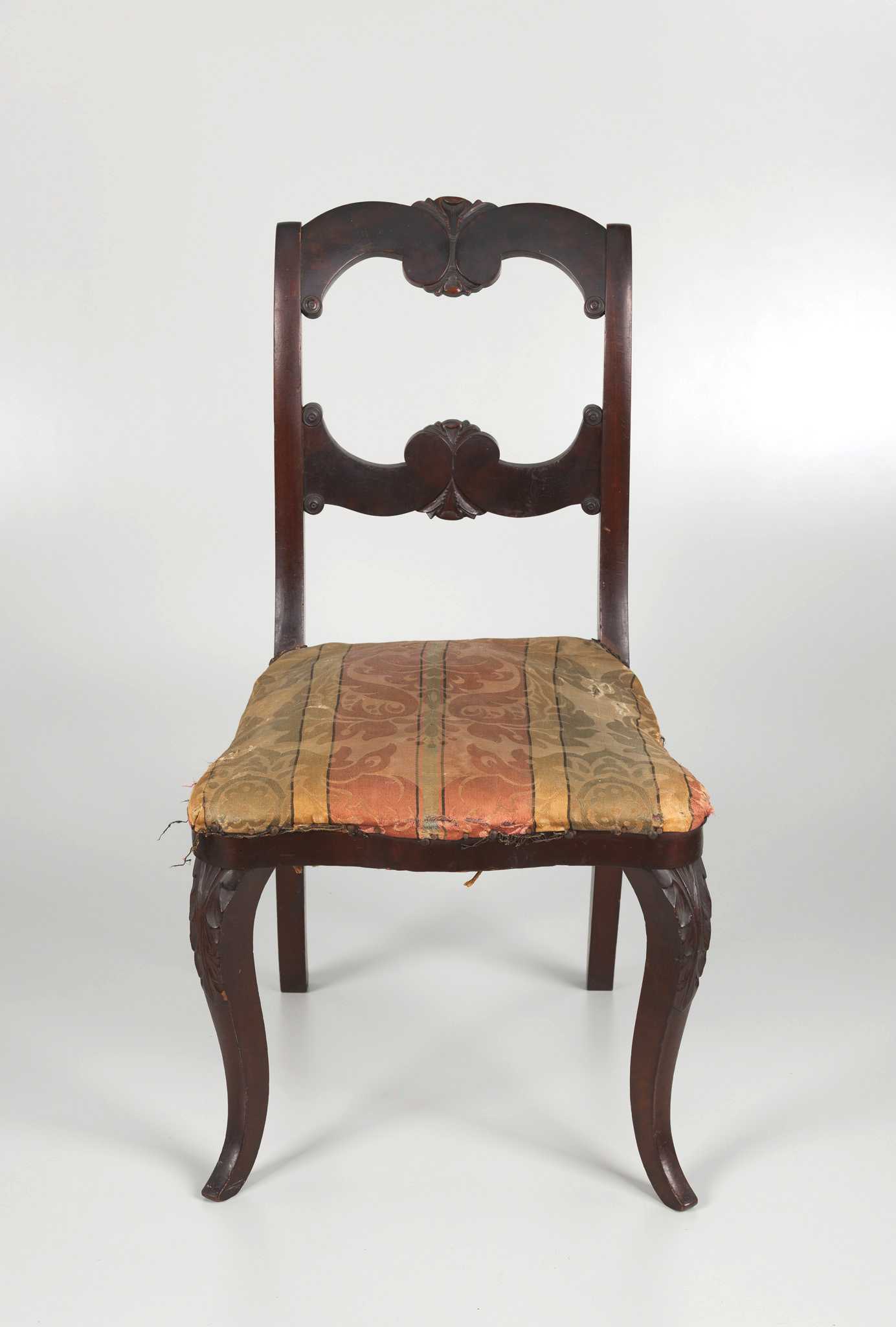 Side chair created by Thomas Day. The chair has mahogany legs and back with scroll and floral detailing. There is an engraved floral motif on the center top rail. The seat is upholstered and filled with burlap. The fabric has cream, green, and red striped sections with black borders and an overall floral motif. The upholstery is worn with small areas of loss. The chairs springs are visible through the loose fabric backing below the seat.
