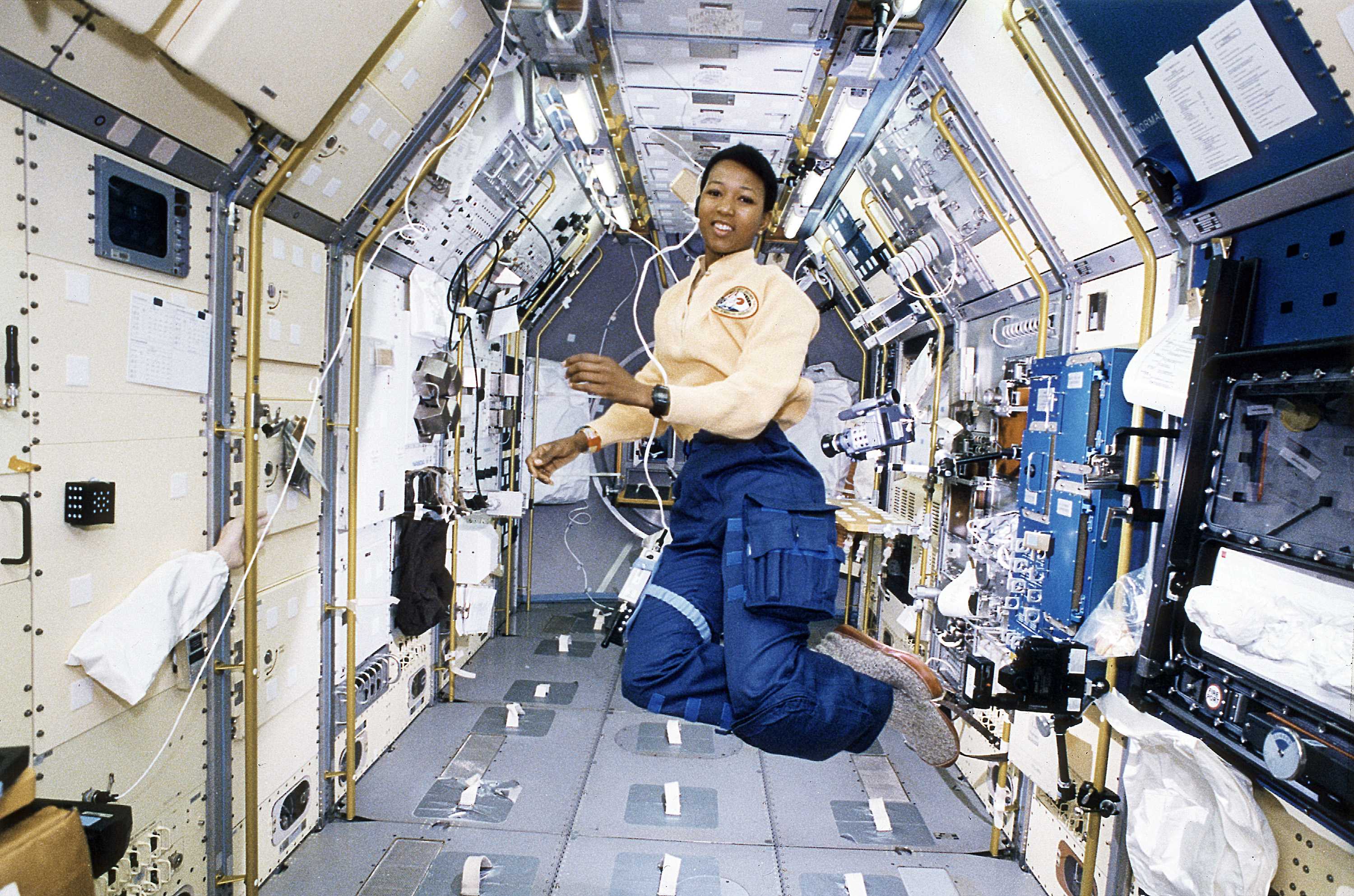 Astronaut Mae Jemison is onboard and floating inside a space craft. She is wearing a blue pants.