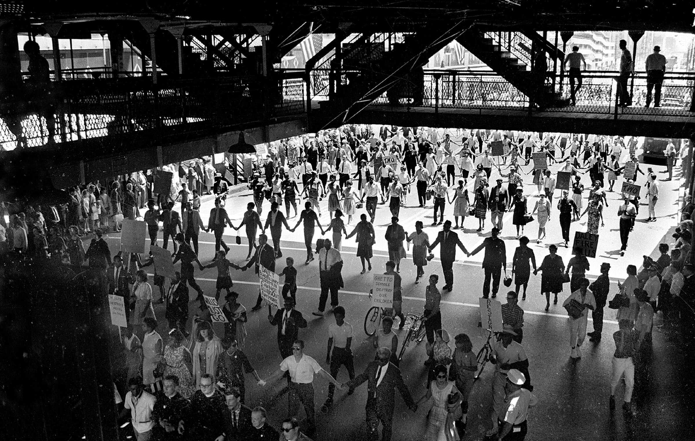 Black adn white photograph of a large group of people holding hands, row by row under a metal platform.  The people are holding signs protesting segregation.