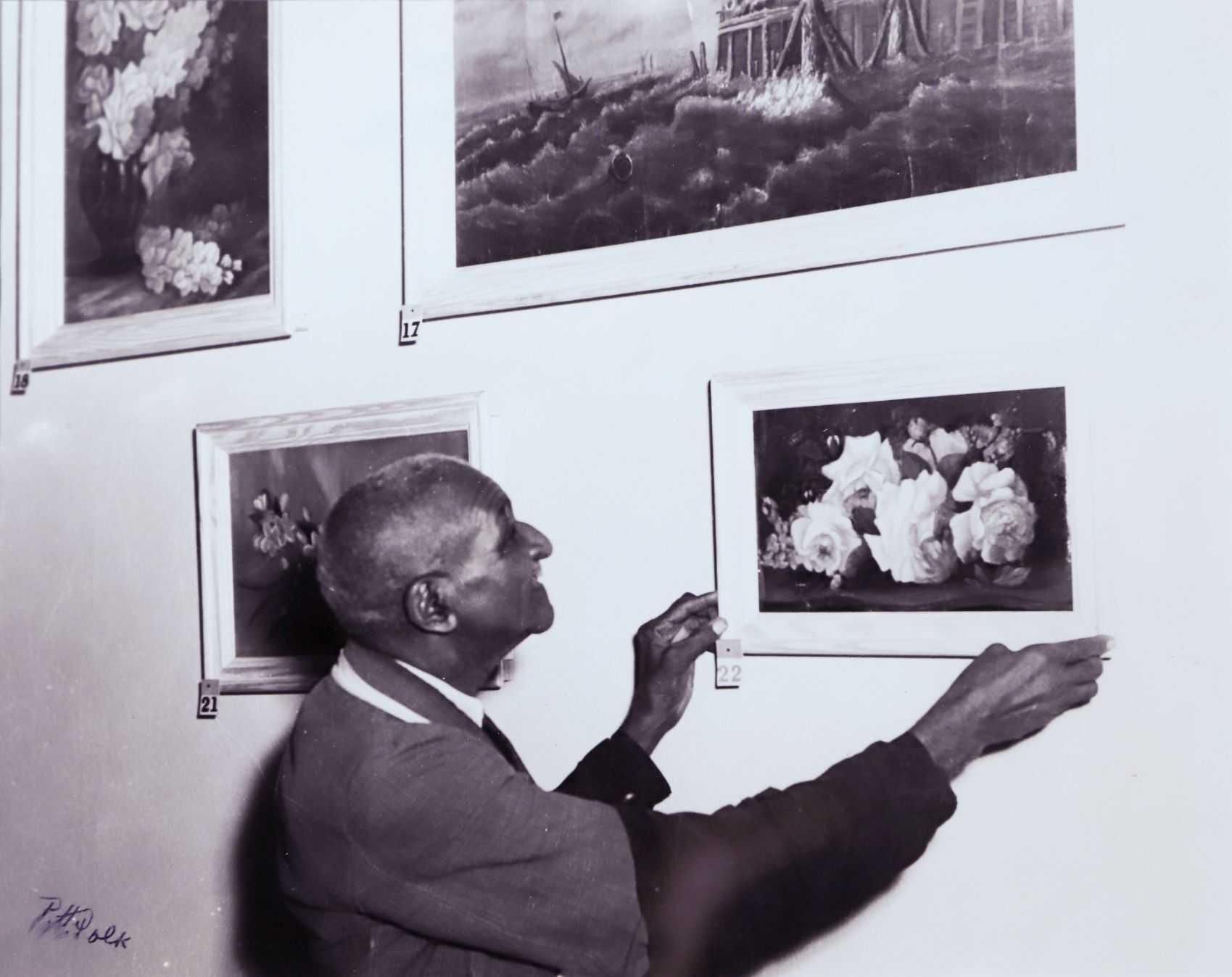 Black and white photograph of George Washington Carver adjusting one of his paintings on a gallery wall.