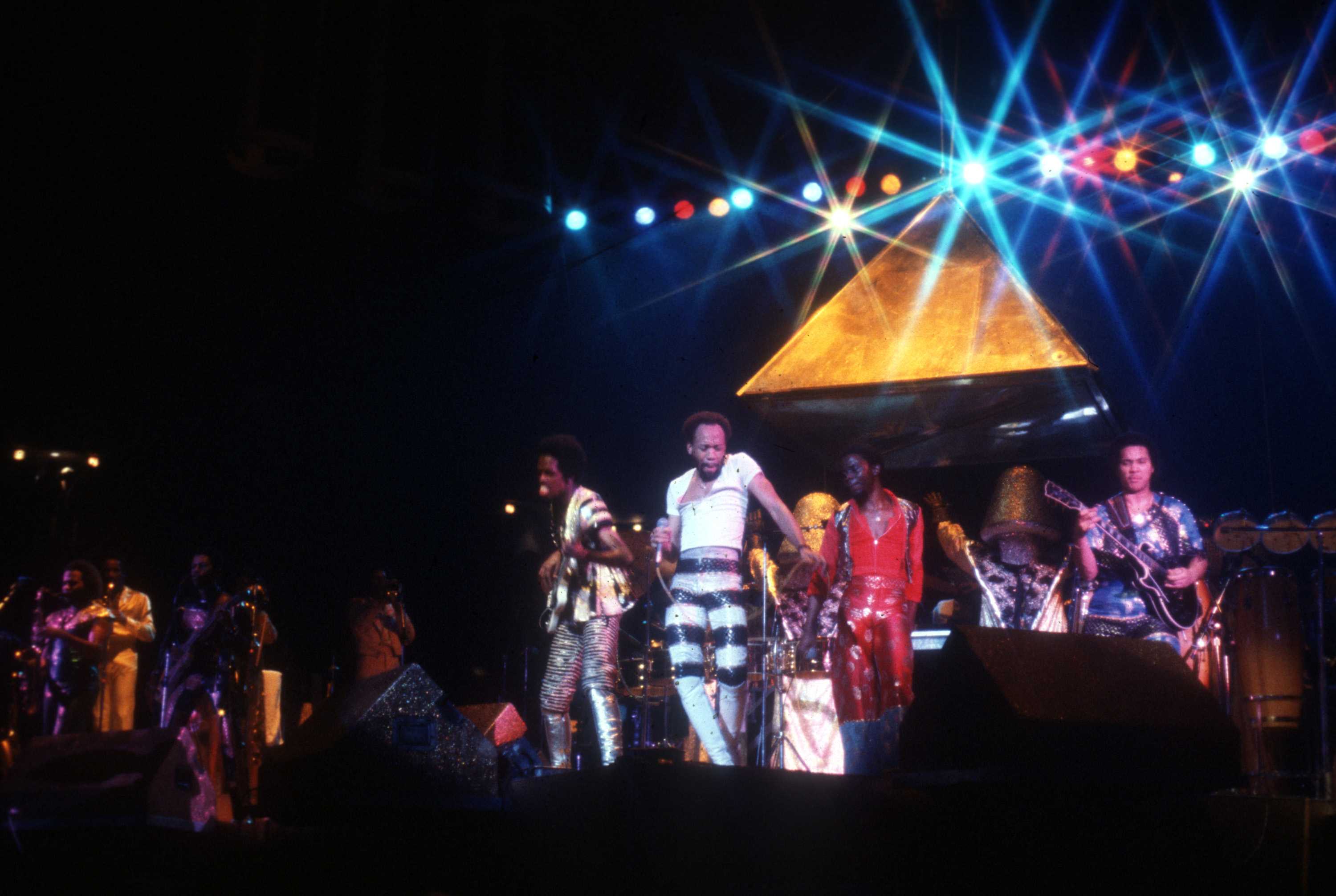 A colored photo of Earth Wind & Fire playing on stage. A gold structure is behind the band as they play.