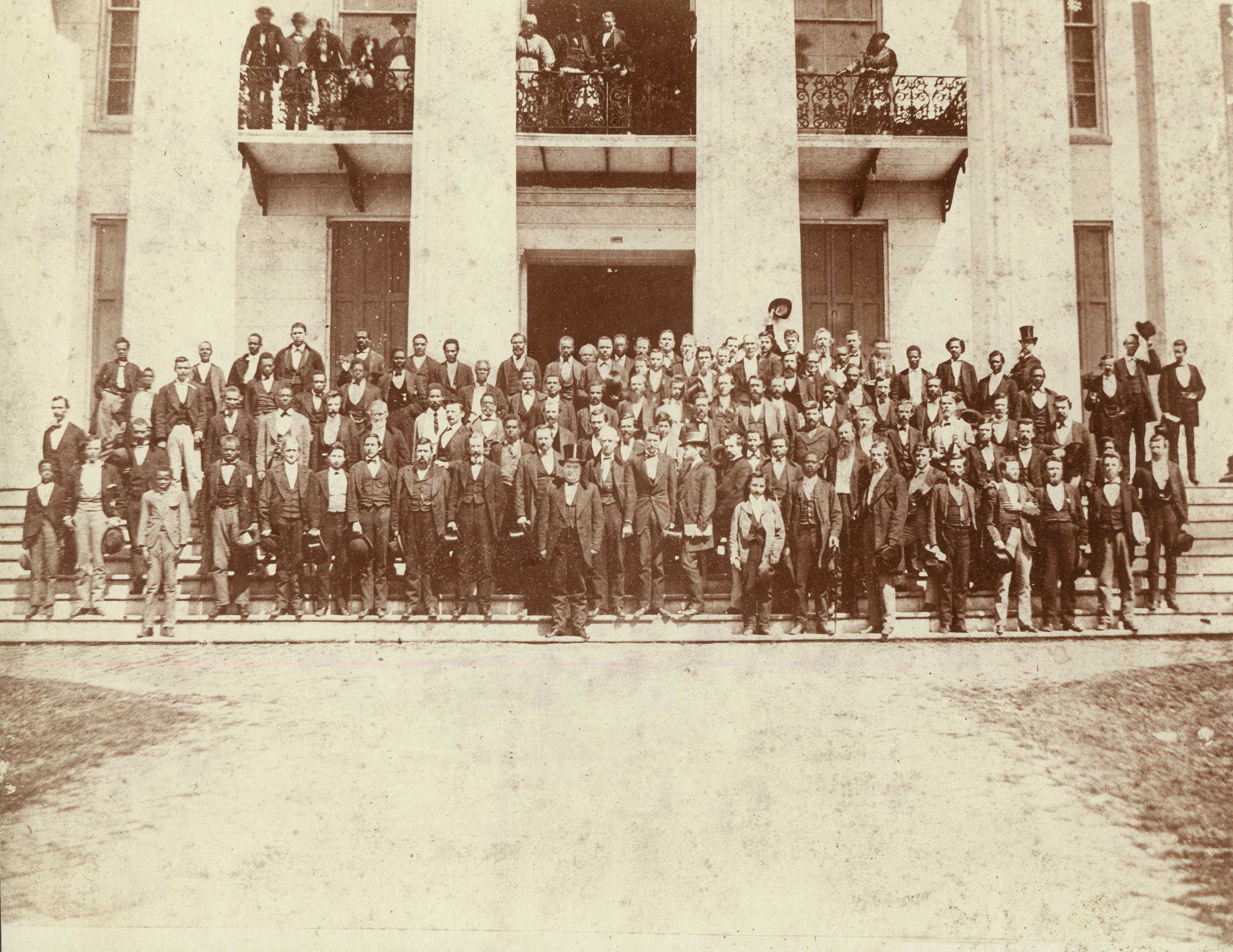A sepia toned photograph of a large crowd of representatives stand in front of the Alabama's state capitol.