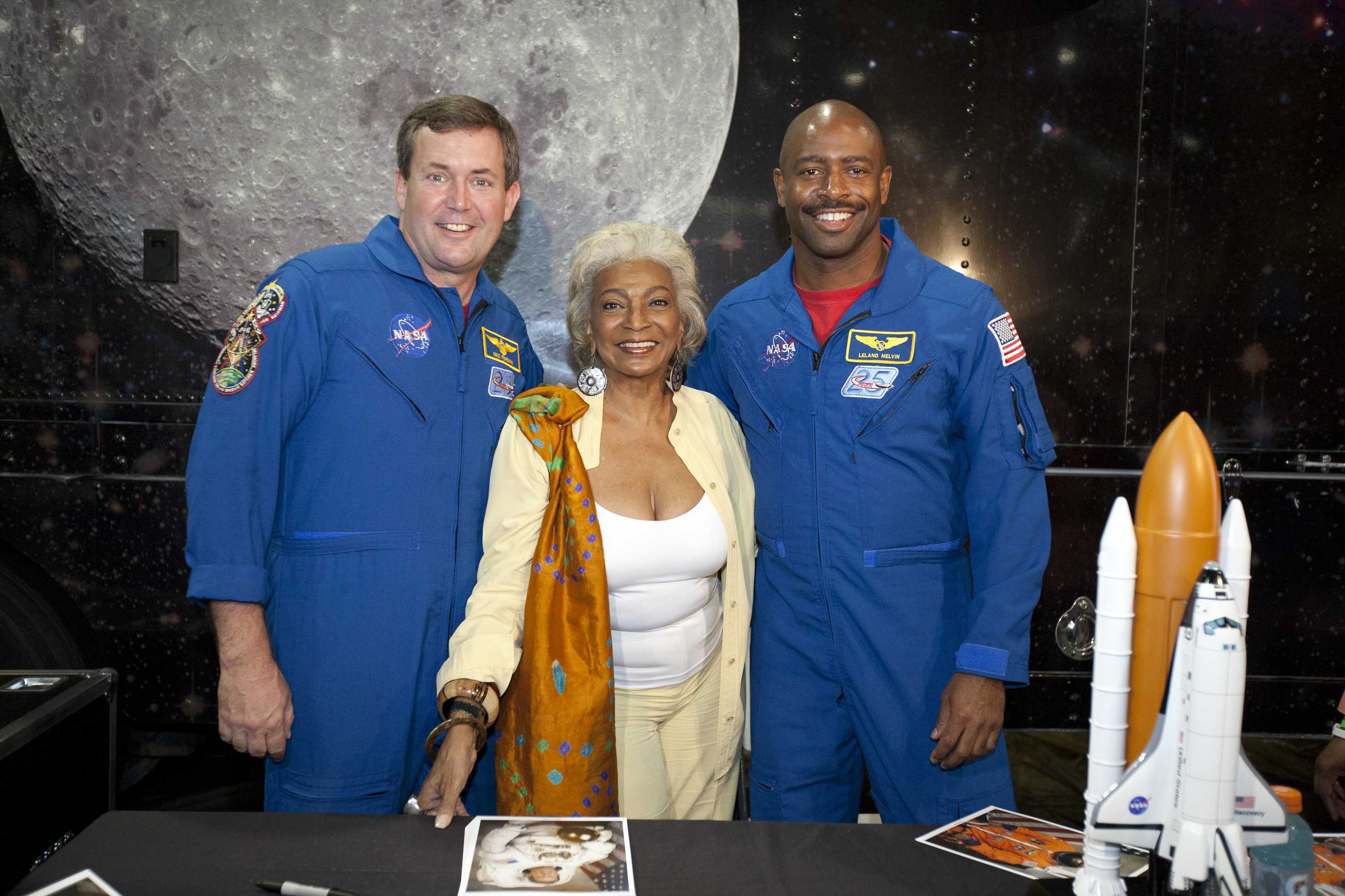Nichelle Nichols smiling and posing for a photo in between two astronauts in front of picture of the moon.