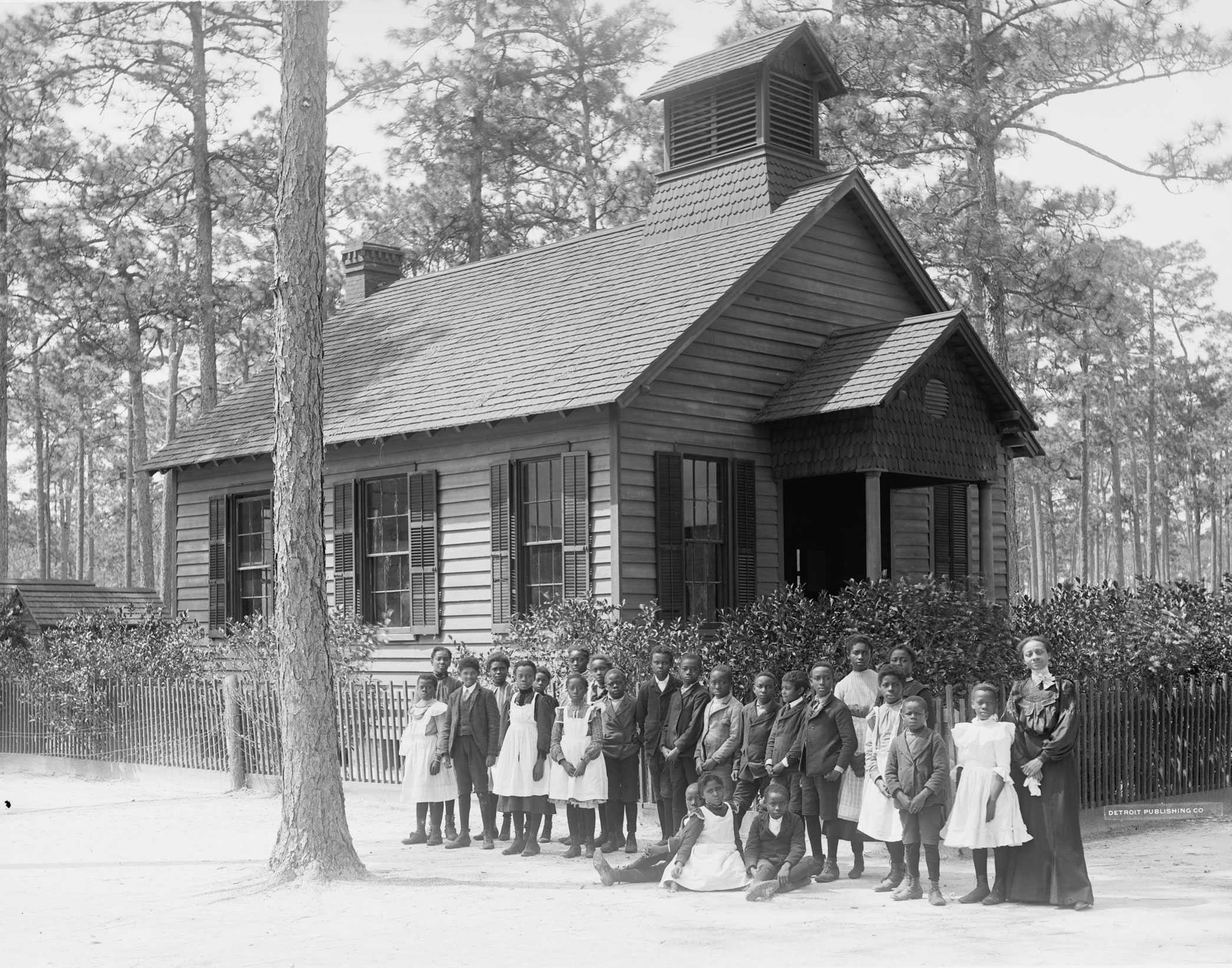 A black and white portrait of a teacher and 20 students standing in front of their school in 1900.