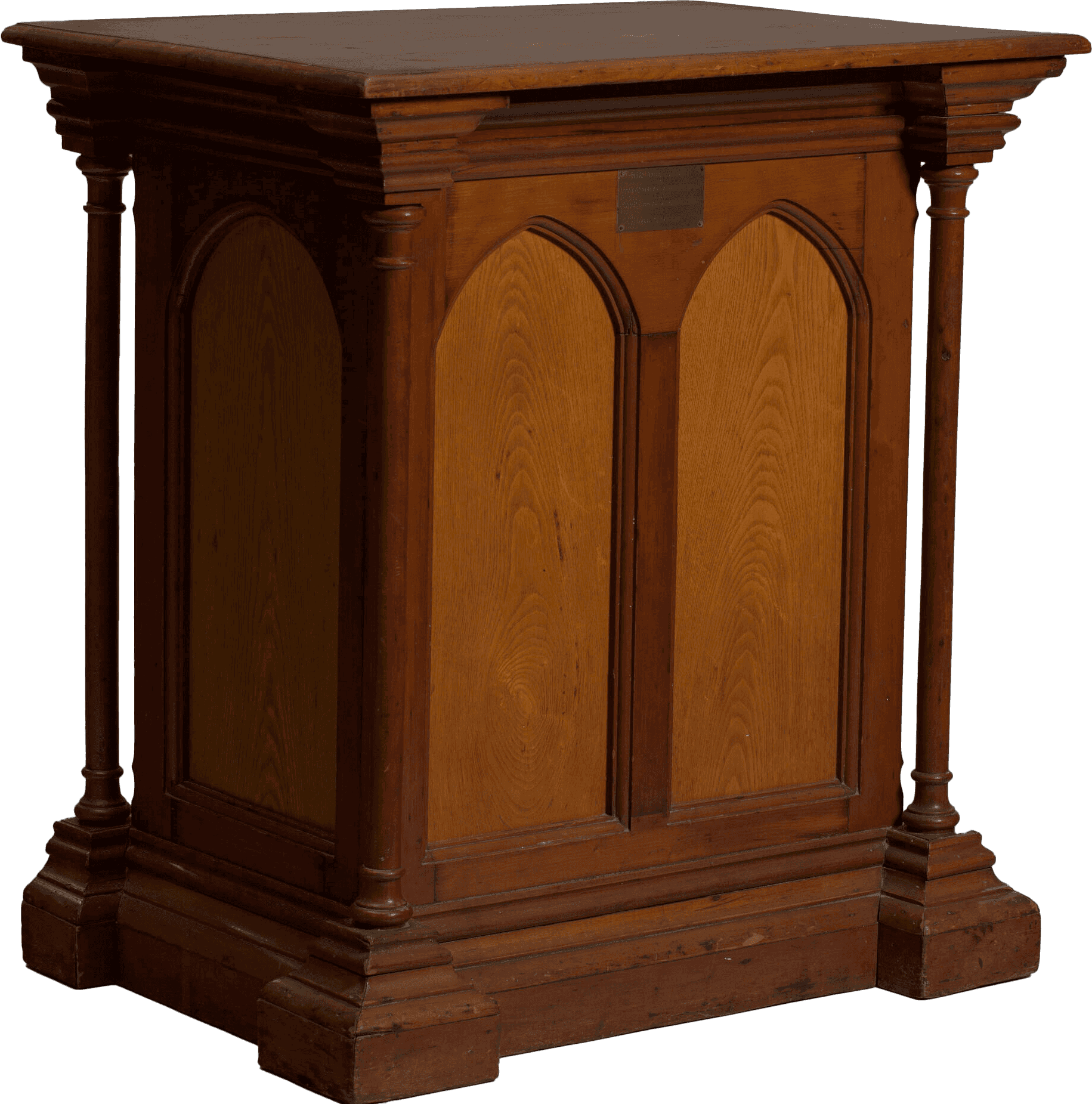 A pulpit from the Metropolitan AME Church, consisting of a hollow, three-sided structure composed of multiple pieces of wood that show different techniques, ages, and varnishes. The front of the pulpit features two arches separated by a piece of wood and framed by thin columns on either side. The wood is smooth and varnished with a light resin through which the wood grain is still visible. The cornices at the top and bottom of the columns are similar in style, with those on the bottom heavier due to extra supporting wood. There are small juts of wood in the interior suggesting a shelf may have been in place inside the pulpit for holding objects. A small metal placard screwed to the front of the pulpit notes “This podium was built for Metropolitan AME Church formerly Union Bethel AME Church by John Simms 1838.”