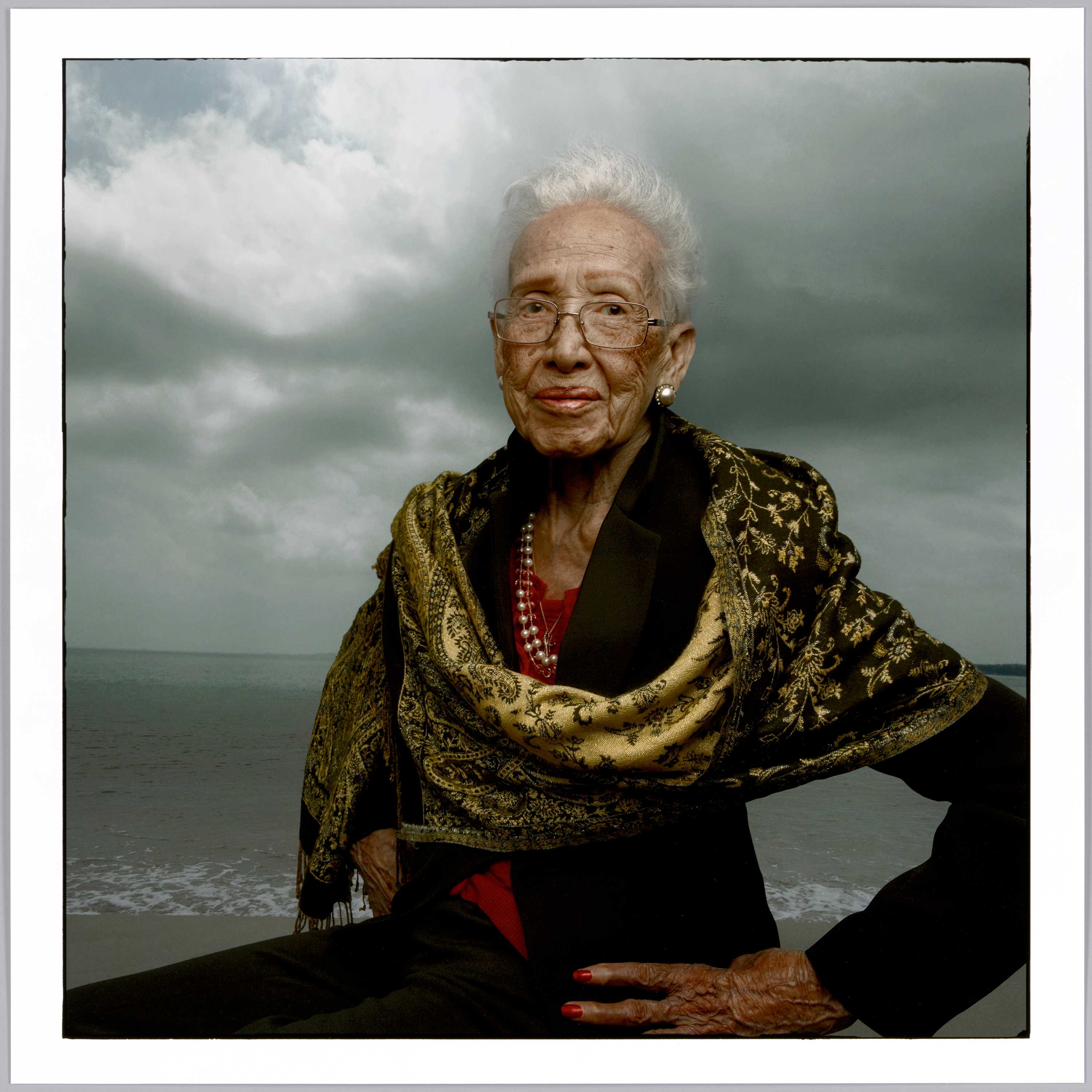 A portrait of Katherine Johnson looking at the camera with a hand on her hip in front of the ocean.