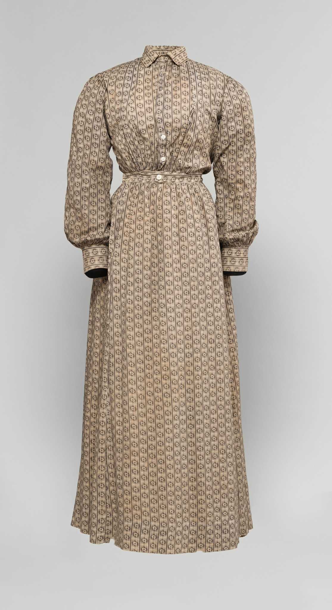 A single piece handsewn dress believed to have been worn by the formerly enslaved woman Tempy Ruby Bryant and handed down through her descendants. The dress is composed of a brown and taupe colored floral patterned fabric. The pattern has columns of flowers bordered by dark vertical lines with light vertical lines running through the flowers. The dress has white buttons at the waist, in a single row from waist to collar. Four buttons are missing: one at the waist and three near the collar. There is neat tucking at the waist band and sleeve cuffs.