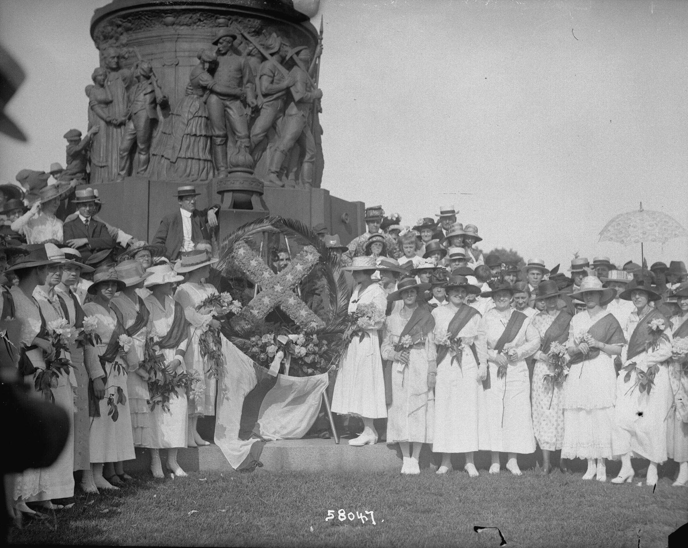 A photo of a large group of The United Daughters of the Confederacy posing together stand together in front of a monument.