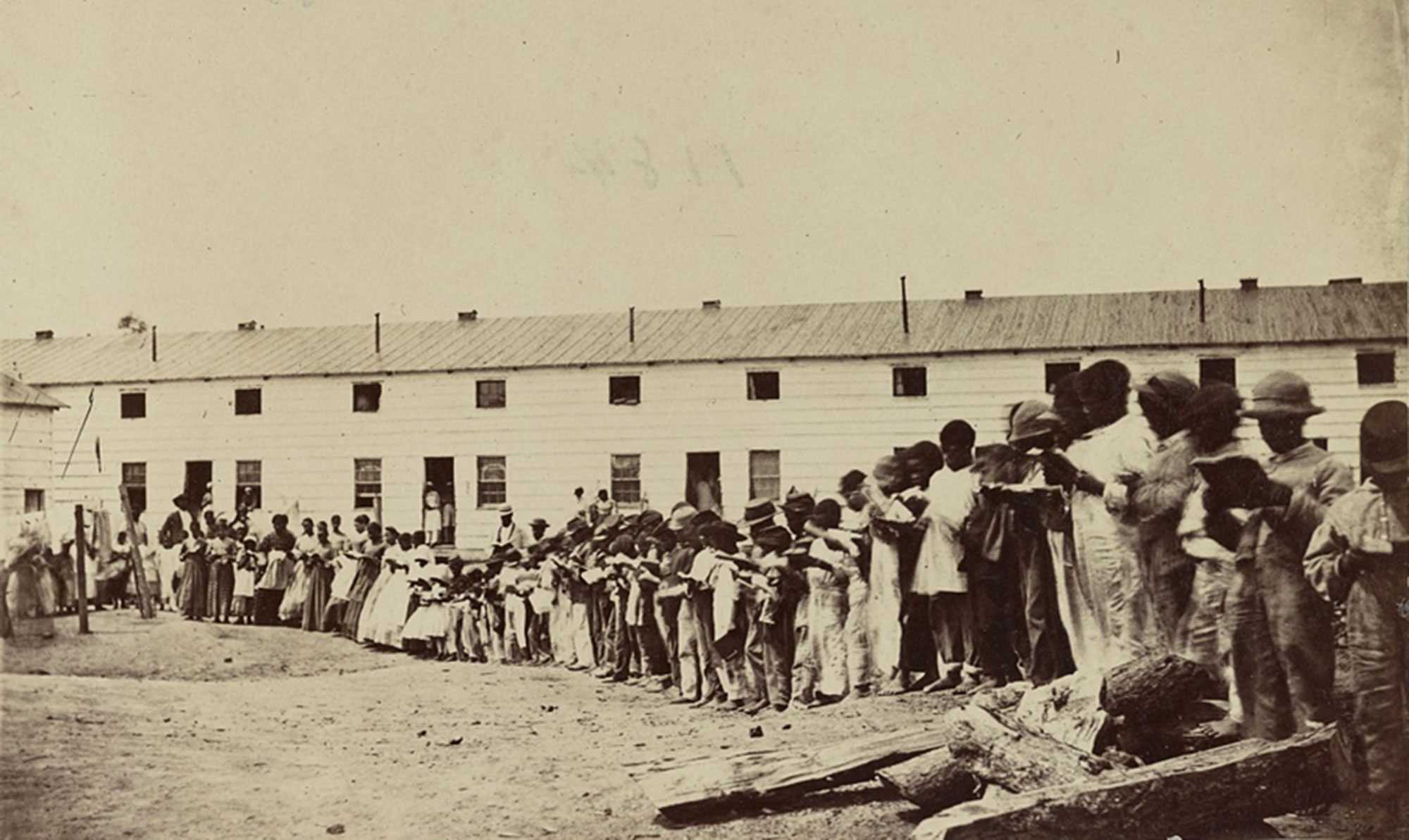 Historic photo of a large group of African American people, including children, standing in a line outside a long building.