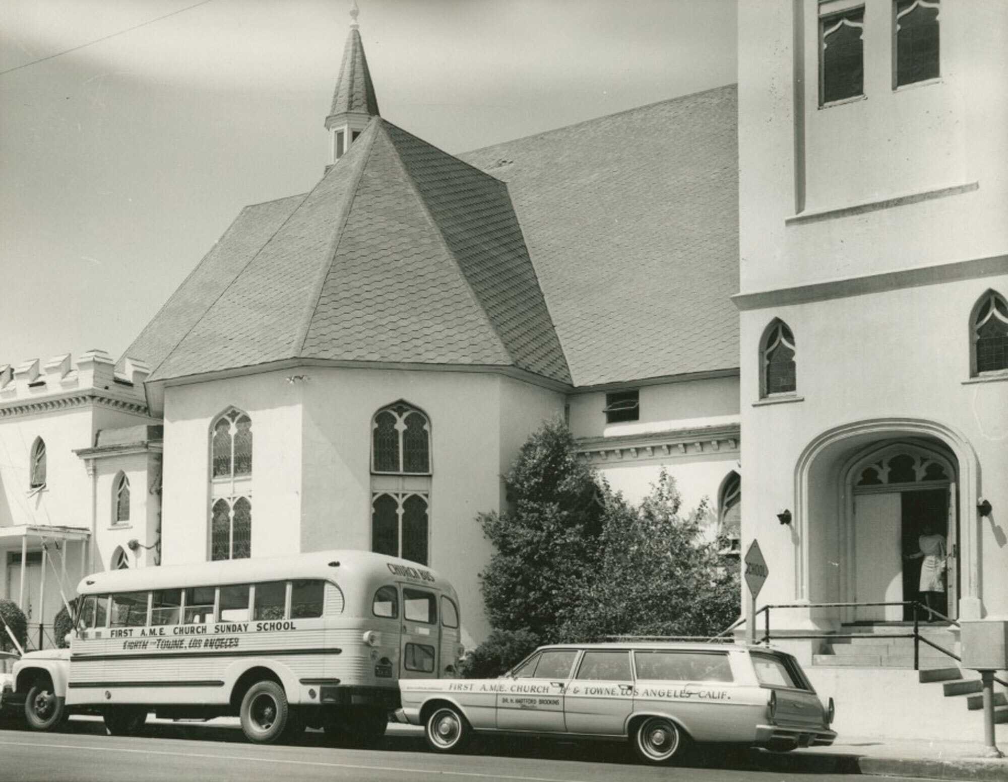 Black and white photograph of a church as seen from the street. The church has a white exterior with gothic style windows of stained glass. On the left half of the image is a multi-walled transept extending from the building. Above this in the background is a cupola. In the foreground in the street on in the bottom left corner is a bus with [FIRST A.M.E. CHURCH SUNDAY SCHOOL/EIGHTH and TOWNE, LOS ANGELES] painted on the side. Behind the bus, right of center, is a sedan with [FIRST A.M.E. CHURCH 8th & TOWNE, LOS ANGELES/DR. H. HARTFORD BROOKINS/Minister] painted on the side. In the lower right corner of the image is an arched and covered doorway with a woman in white emerging. On the reverse of the image is a stamp in the center set at a diagonal [J. BANKS/HOME PORTRAITURE PHOTOGRAPHER].