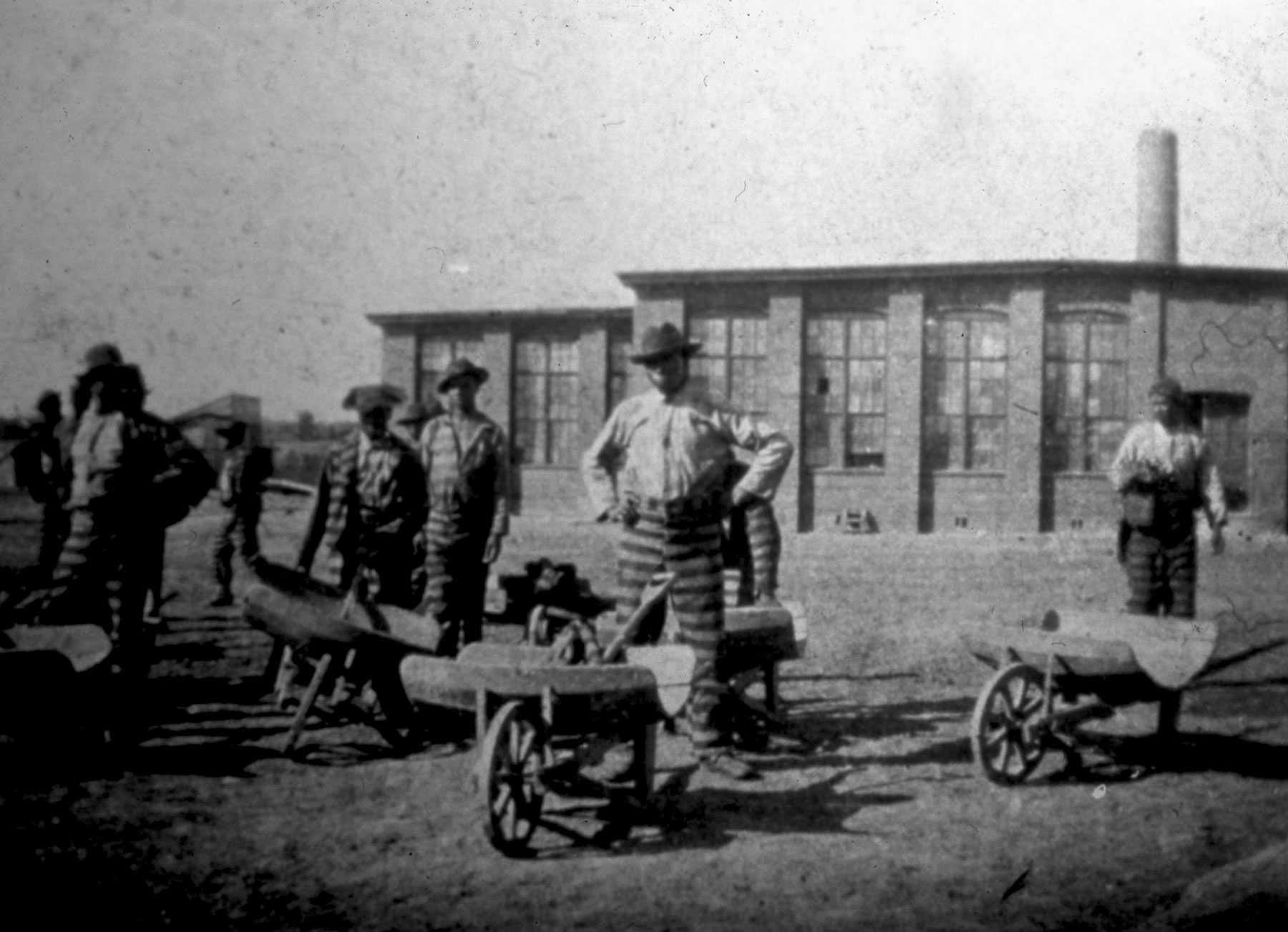 A blurry black and white photo of convicted workers standing with wheelbarrows in front of a factory.