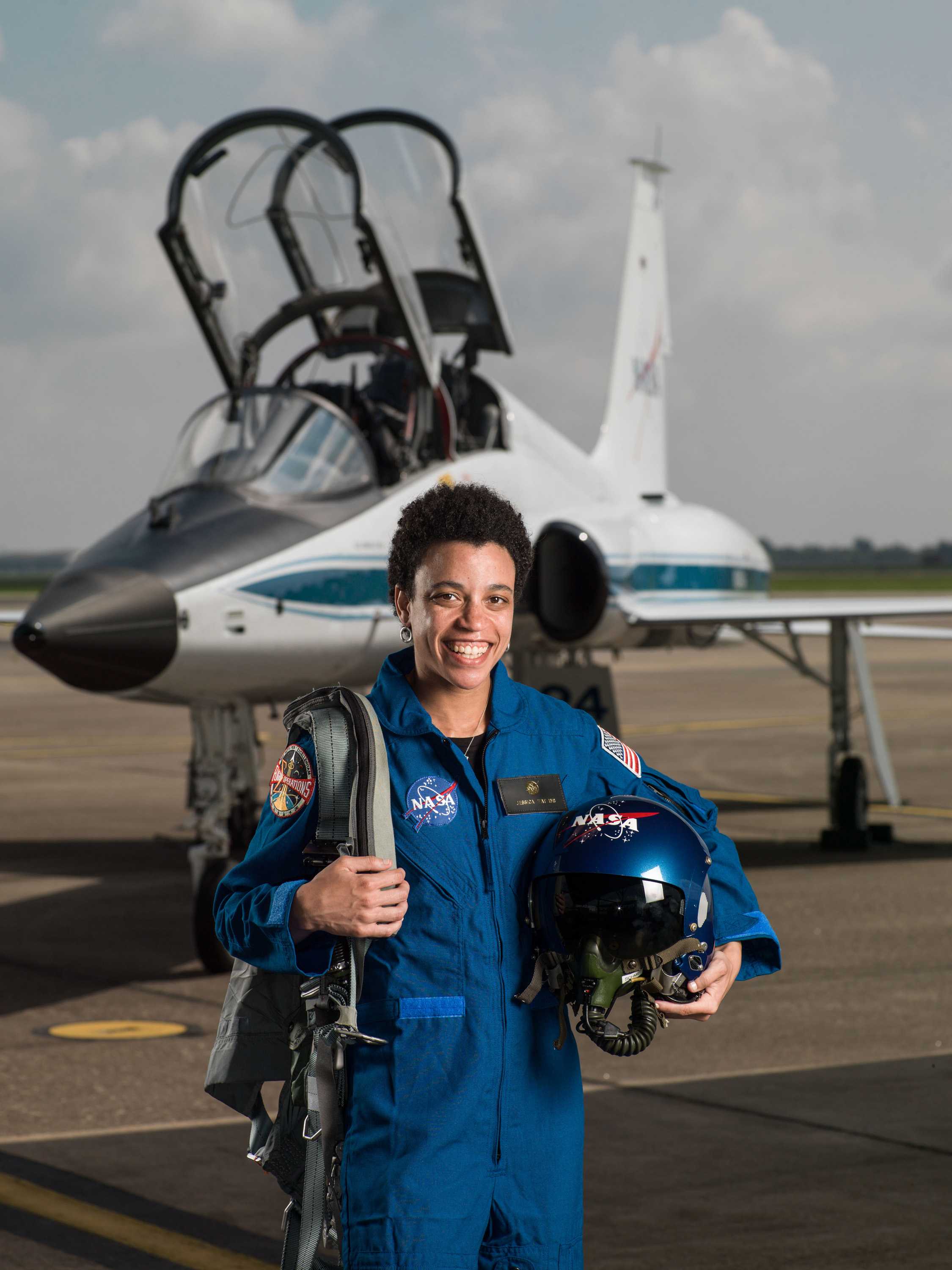 Watkins stands in front of an airplane, posing for a portrait. She is wearing a blue NASA suit and holding a helmet and gear.