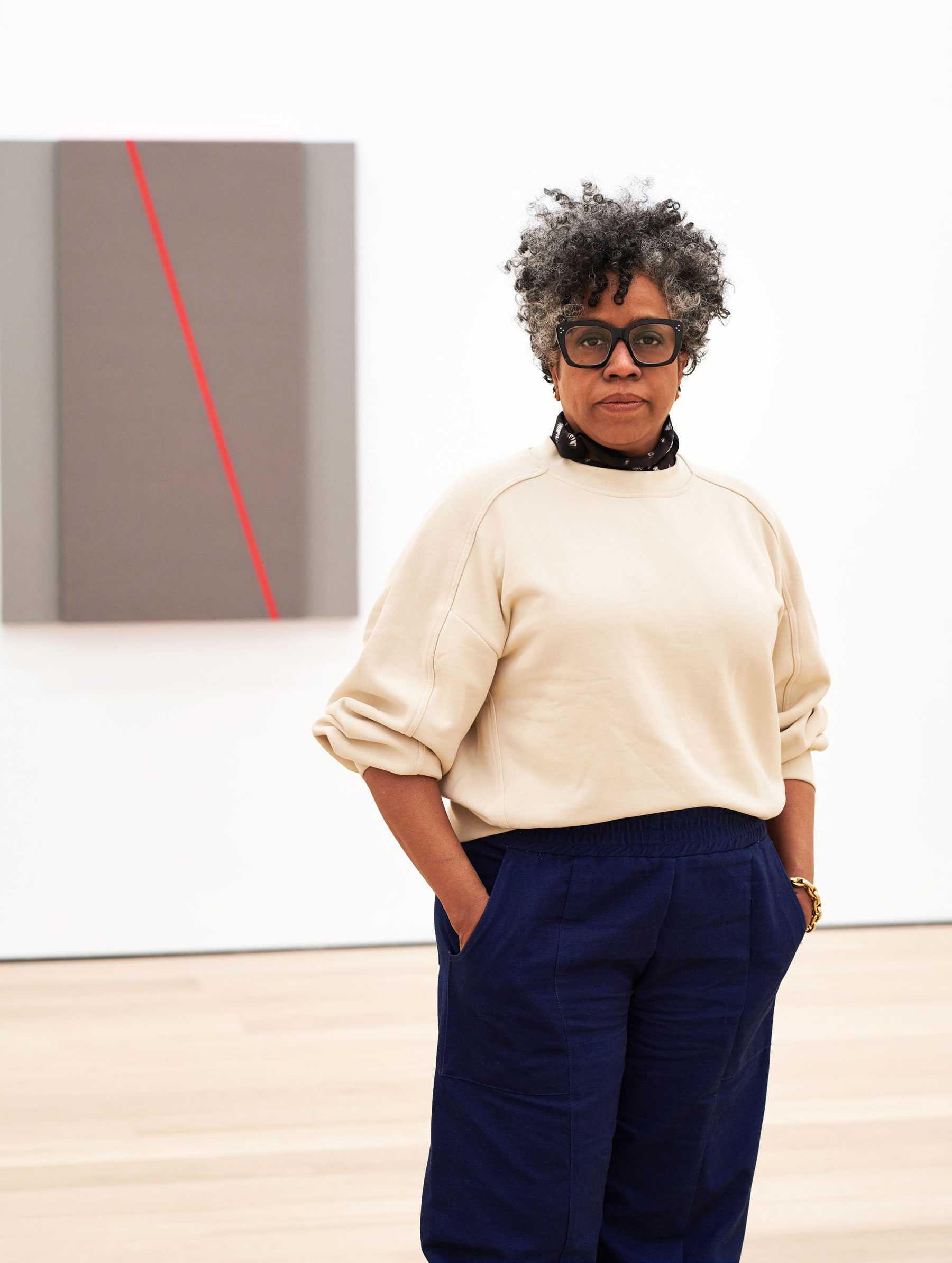 Woman standing in a gallery with abstract art on the wall behind her, hands in pockets, wearing glasses, a beige sweater, and blue pants.