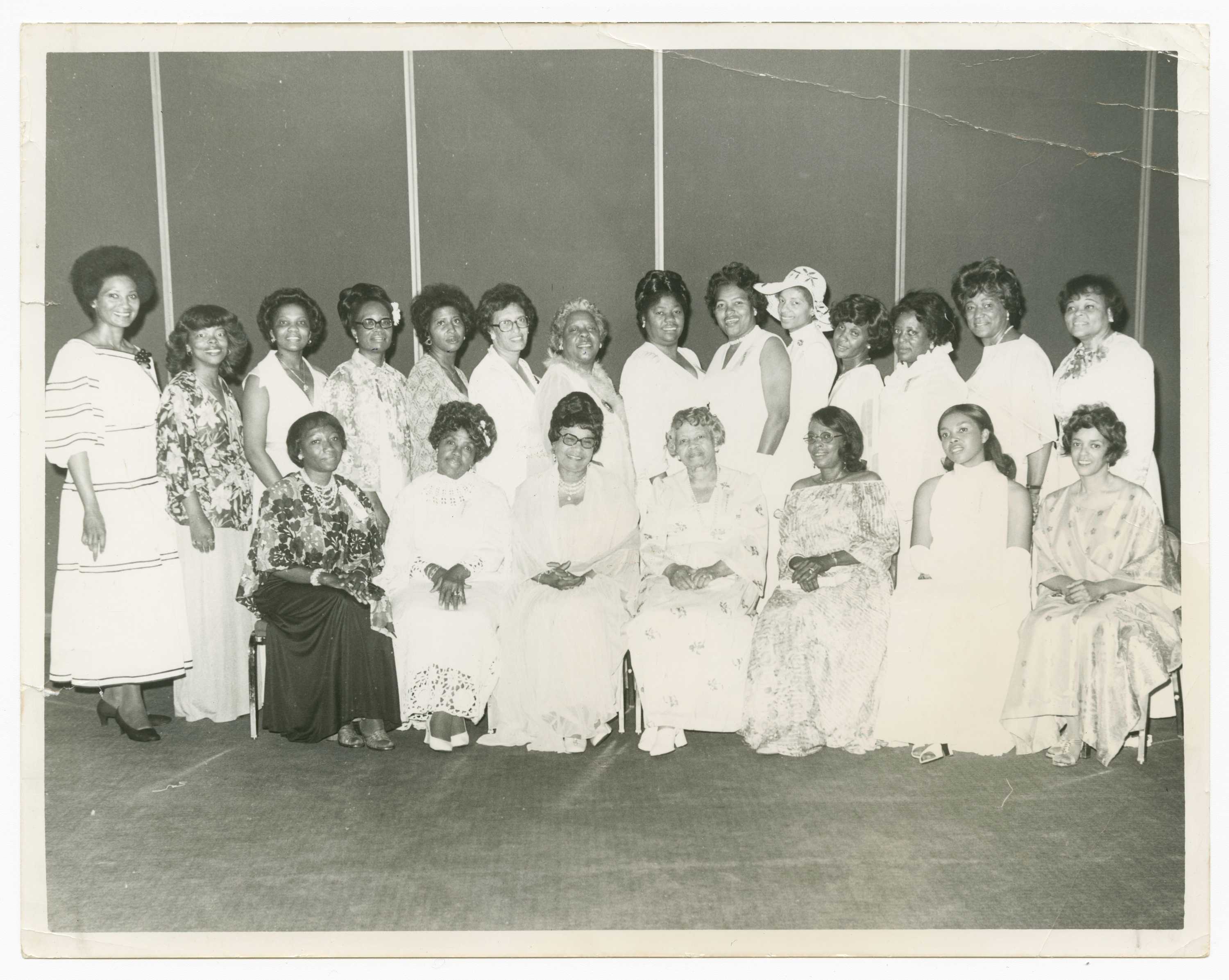 Black and white image of two (2) rows of women. The front row includes seven (7) seated women. The row behind includes fourteen (14) women standing. The women wear a variety of dresses in different styles. Many of the dresses are patterned or with floral prints. One woman wears a hat and stands fifth from the right in the back row. The women are all in a large interior space. On the reverse are a variety of marks. One large angular mark takes up a large portion of the back from the upper right corner to the lower left corner and down to the lower right corner. Along the top half of the back is the image caption with additional numbers and notes. [Ushers for FAMEs 100th Anniversary / Sent" / "Church / 7-28-77 / 4Cals/Hostesses]. In the lower right corner are two (2) stamps with the photographer information and the date [PLEASE CREDIT PHOTO / BY / HARRY H. ADAMS / 4300 SO. CENTRAL AVE. / LOS ANGELES, CALIF. 90011/235-2742 / JUN 1977].