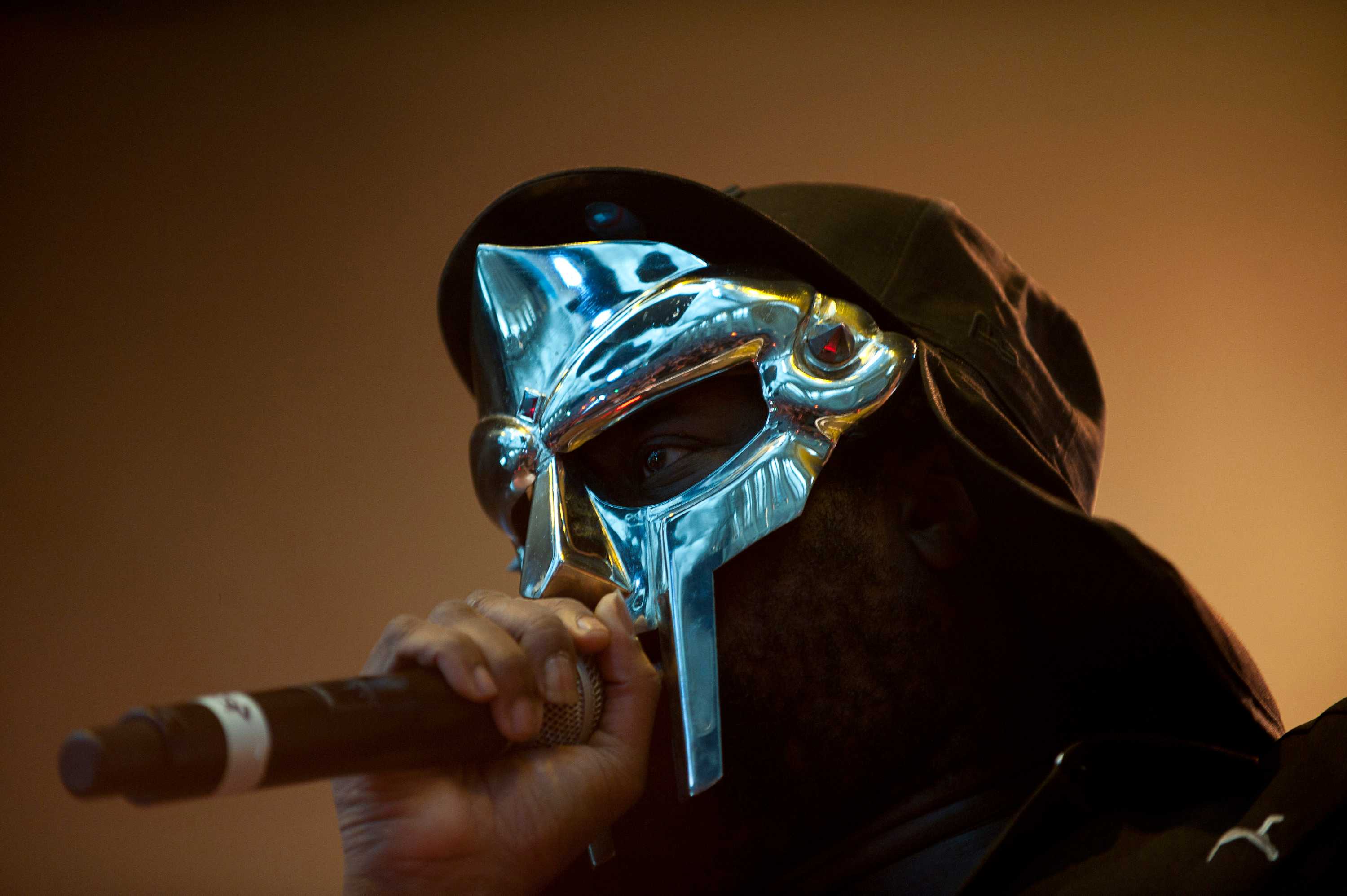 MF DOOM preforming a sliver plated mask that is covering most of his face. He is rapping into a microphone.