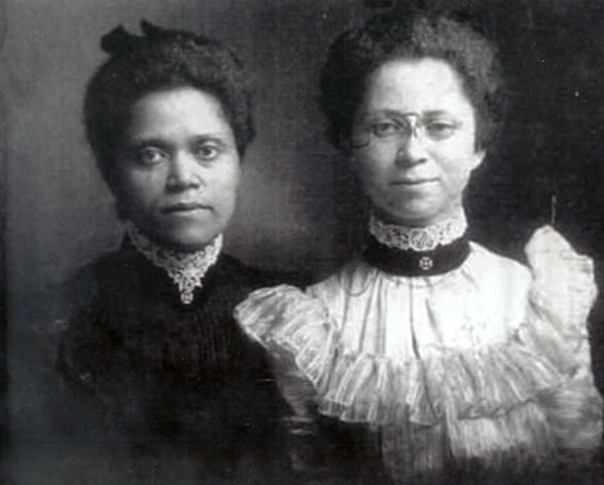 Black-and-white historical photograph of two women standing side by side, dressed in formal late 19th or early 20th-century attire.
