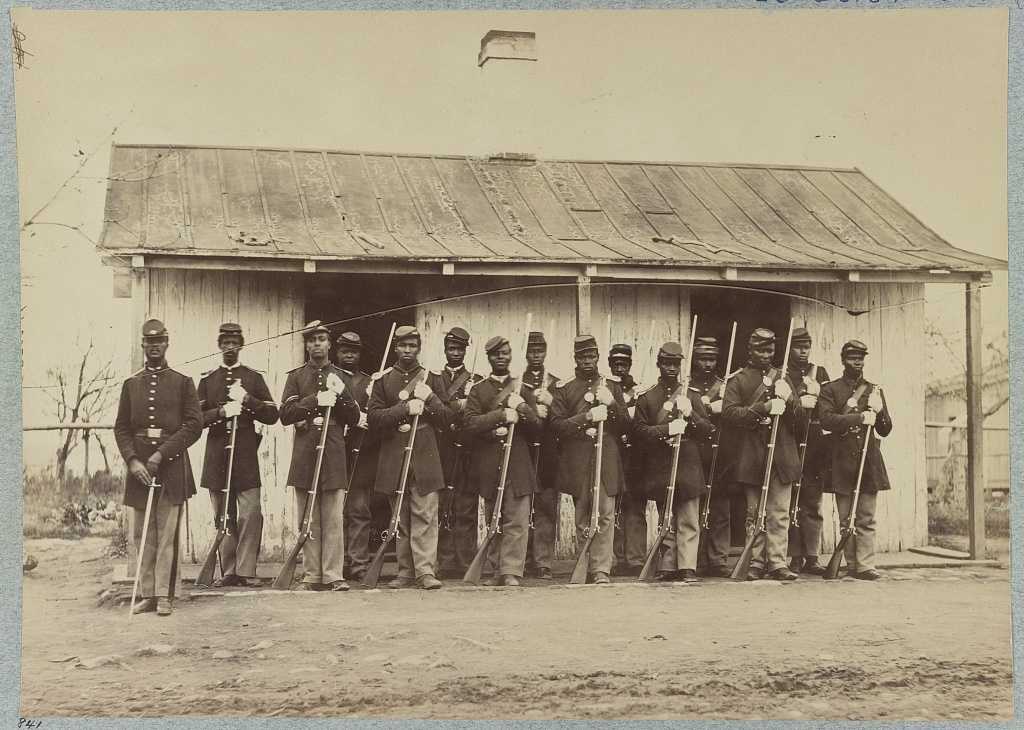 Black Union soliders stand next to each other in front of a small house.
