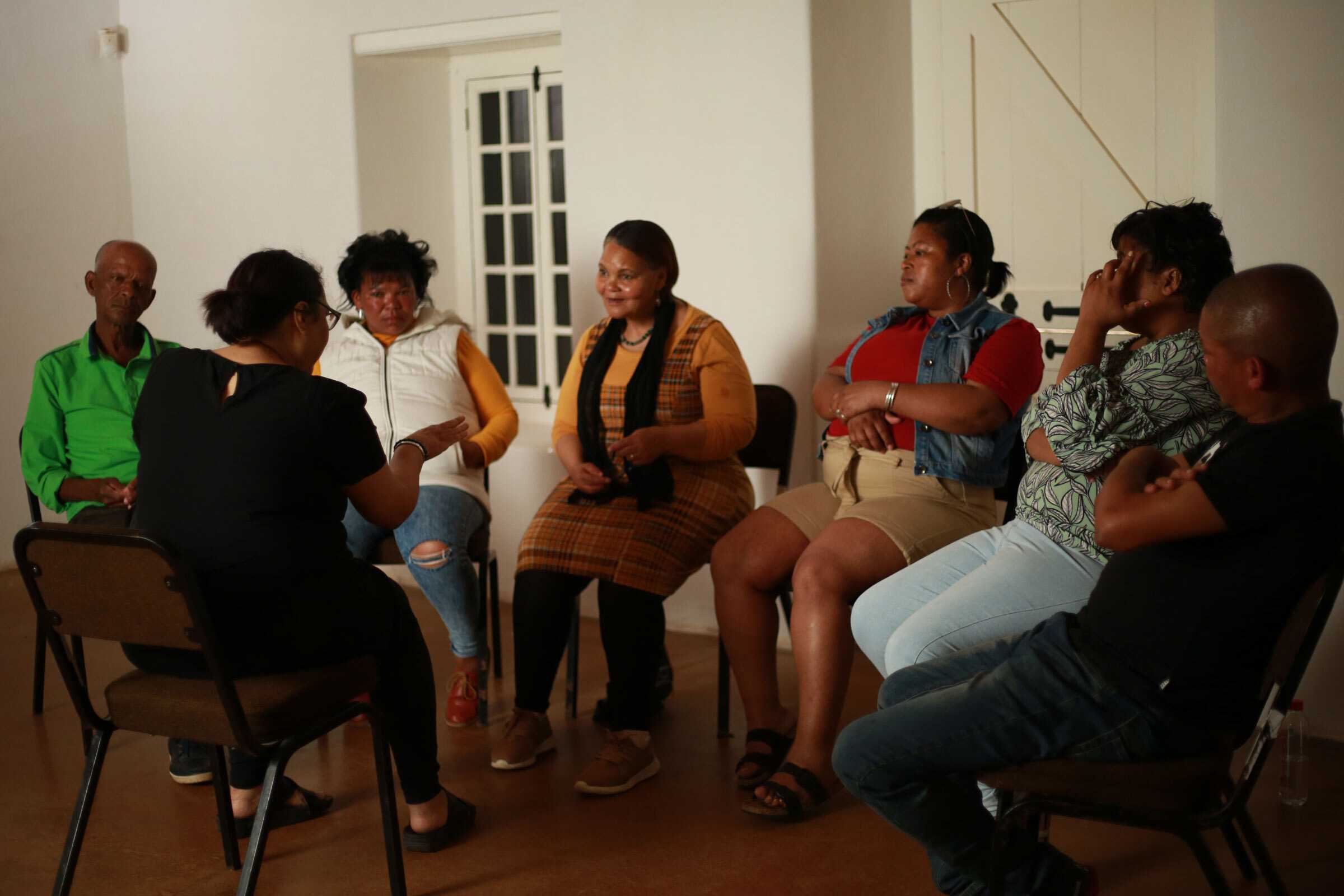 A small group of people seated in a circle of chairs during a discussion or meeting in a bright indoor room.