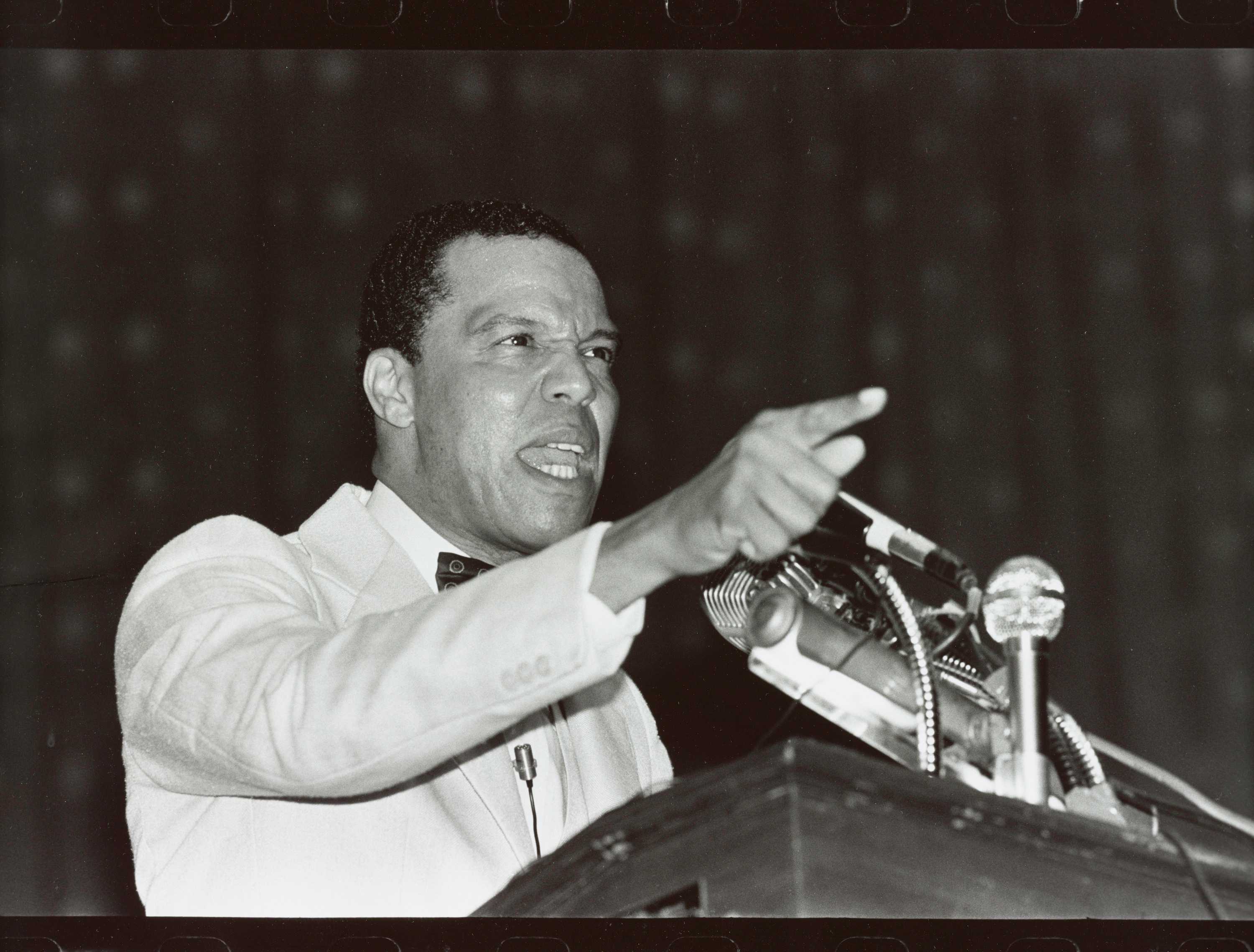 A black-and-white photograph of Louis Farrakhan speaking at a podium and gesturing with a pointed finger. The photograph is stamped and inscribed on the back.