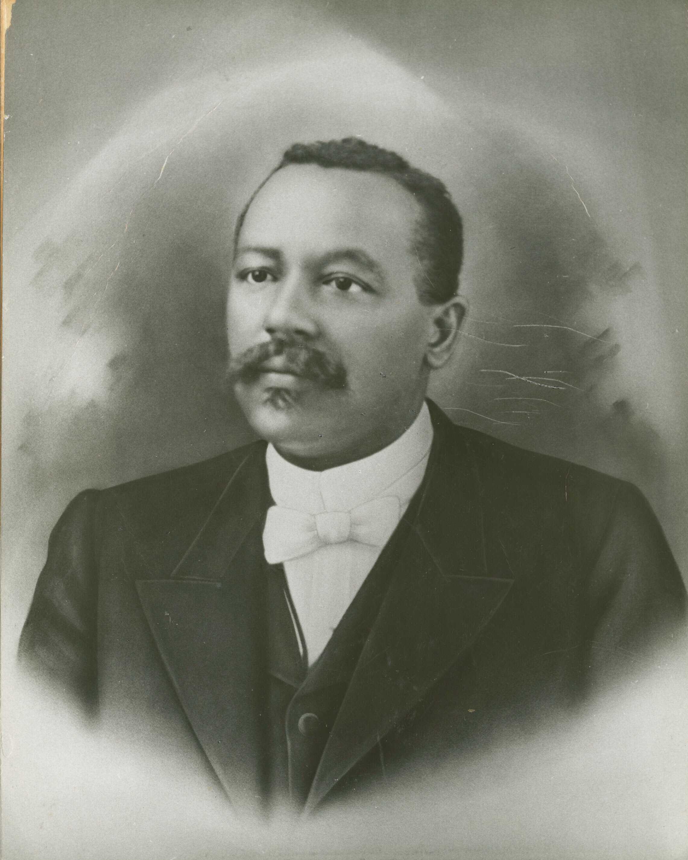 Black and white portrait image of a man wearing a dark jacket, dark vest, white shirt, and white bow-tie. He has a mustache and goatee and looks the left of the camera. The image is hazy at the corners and along the bottom. The background is darker near the man's head. The image shows the man from the chest up in three-quarter view. The photo is adhered via glue to a piece of cardstock the same size as the photo.