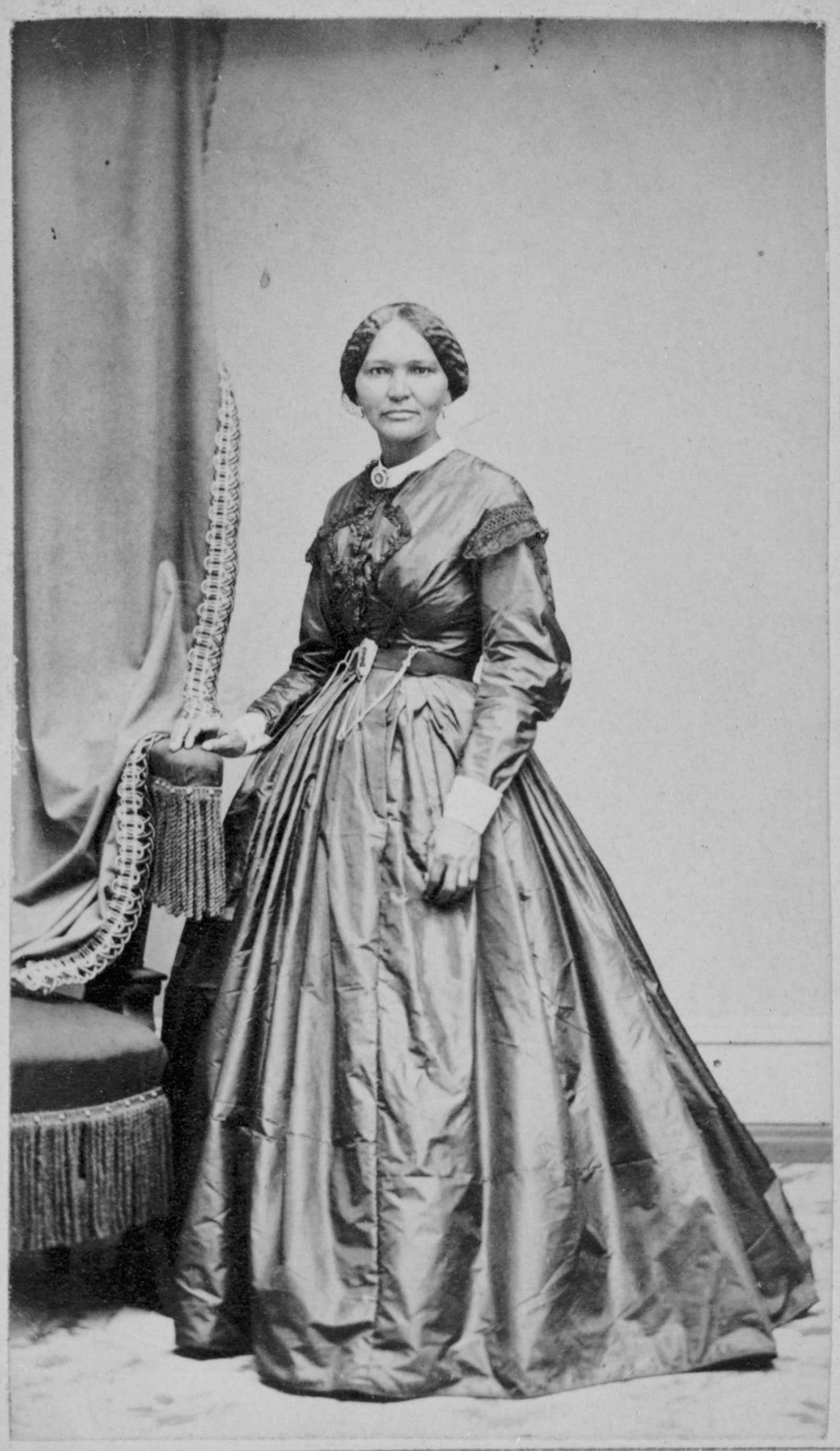Elizabeth Keckly posed for a portrait. She stands in an elaborate gown with her hand resting on a chair.