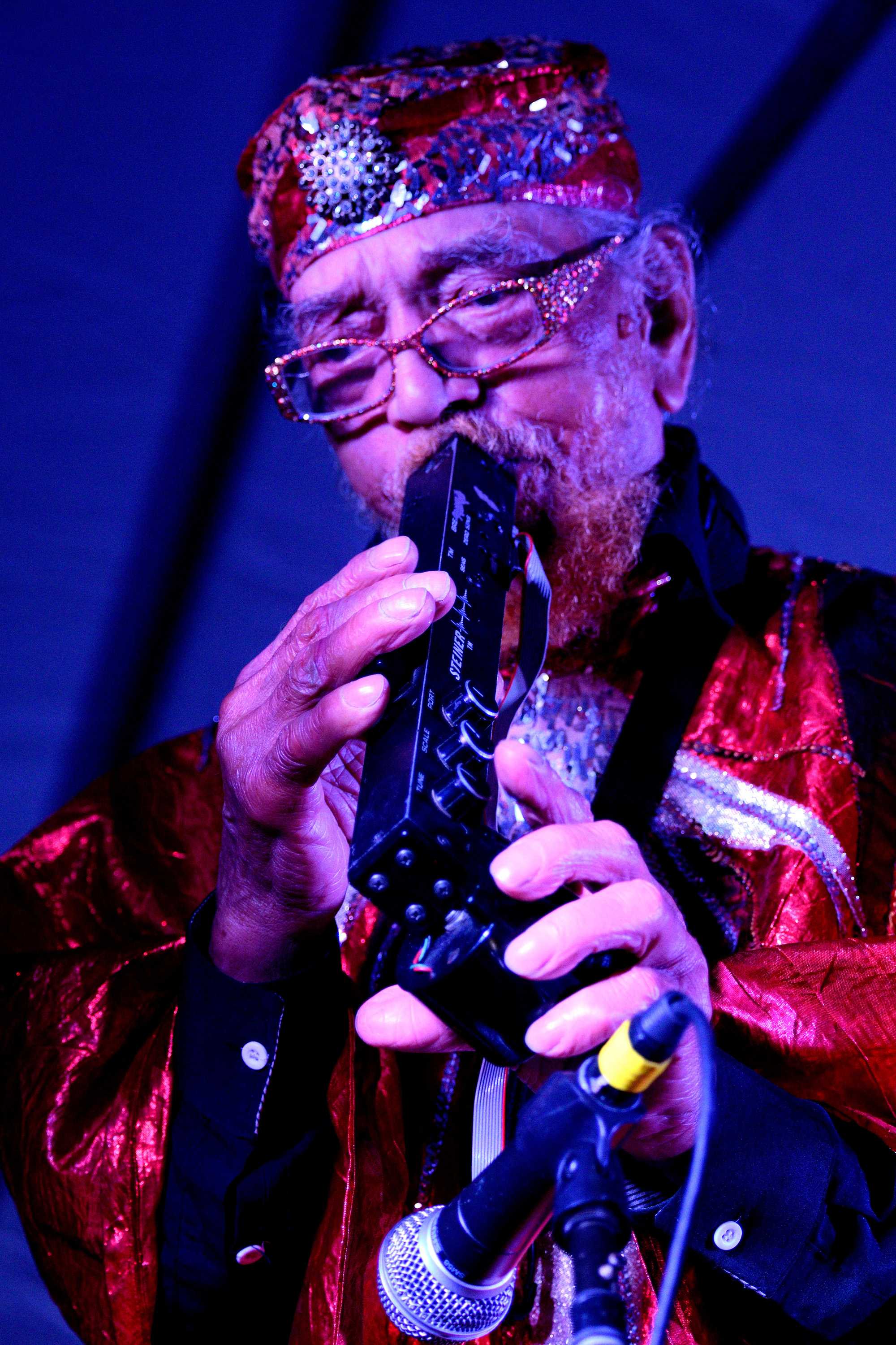 Marshall Allen playing a black saxophone like instrument while dressed in costume and wearing glasses.
