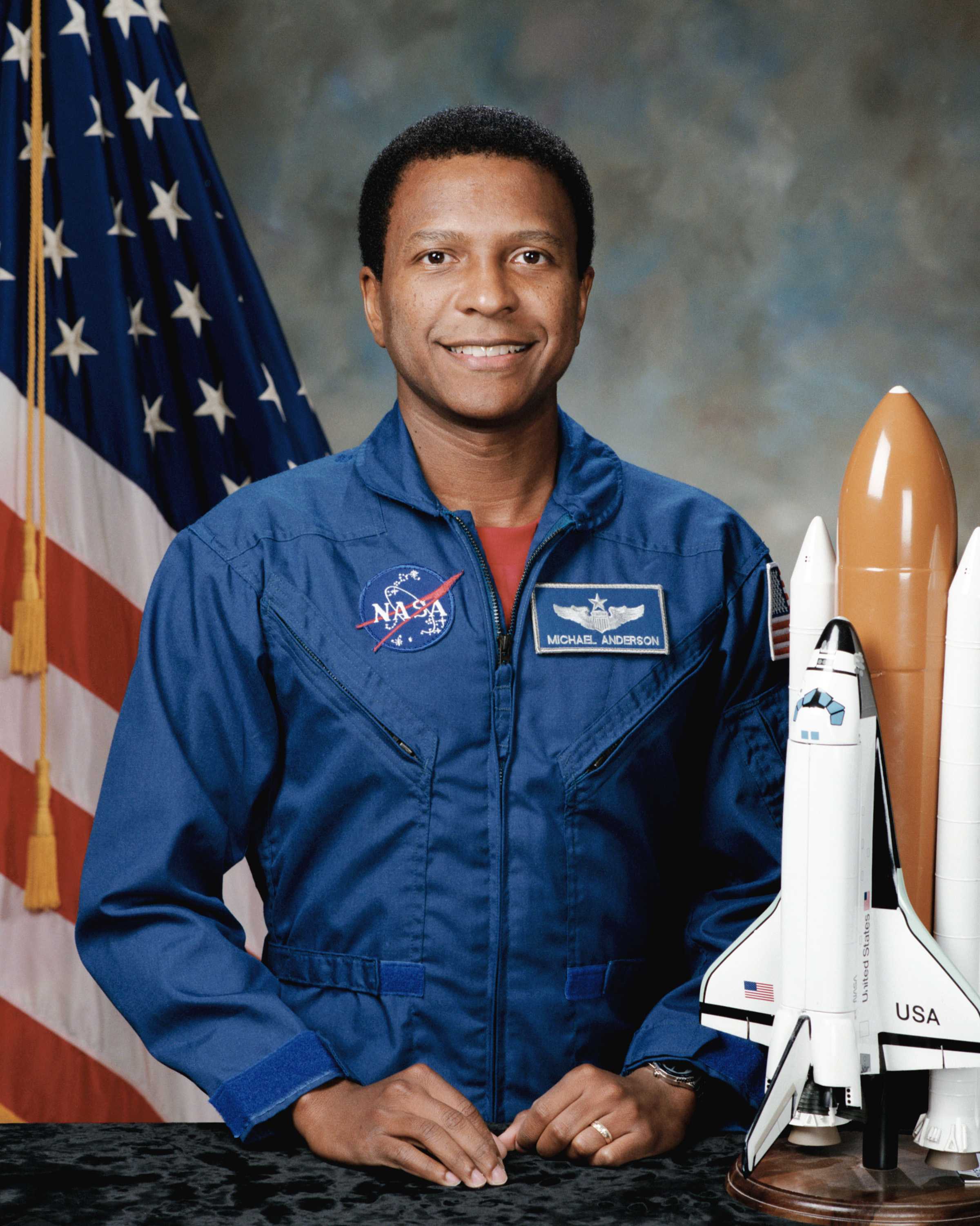 Anderson poses for his portrait while wearing a blue NASA suit in front of a USA flag and model spaceship.