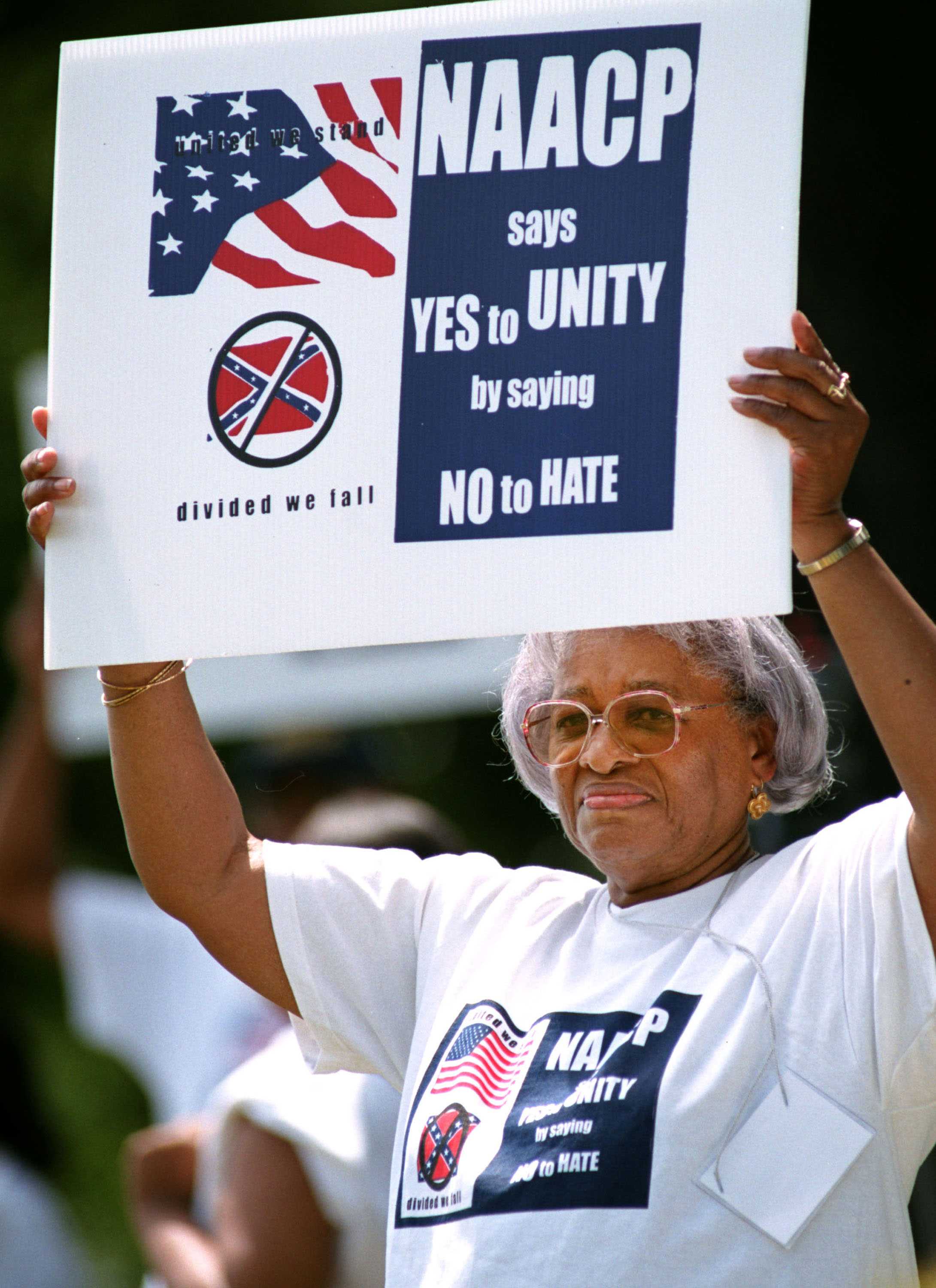 An older black woman is holding up a sign that says 'NAACP says Yes to Unity by saying No to Hate'