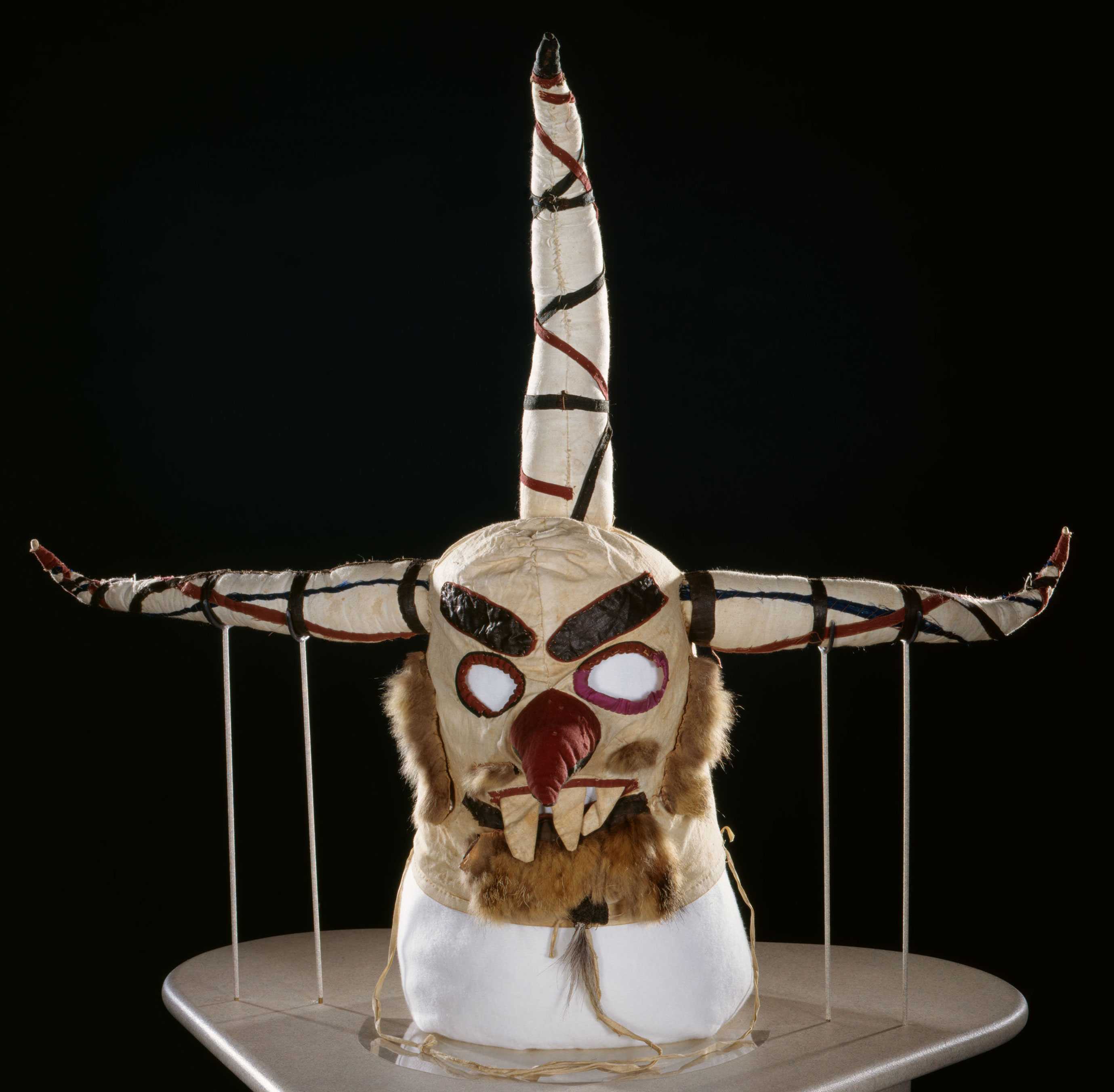 Face mask with red eyes and nose and three horns decorated in black and red.  There is fur representing facial hair.