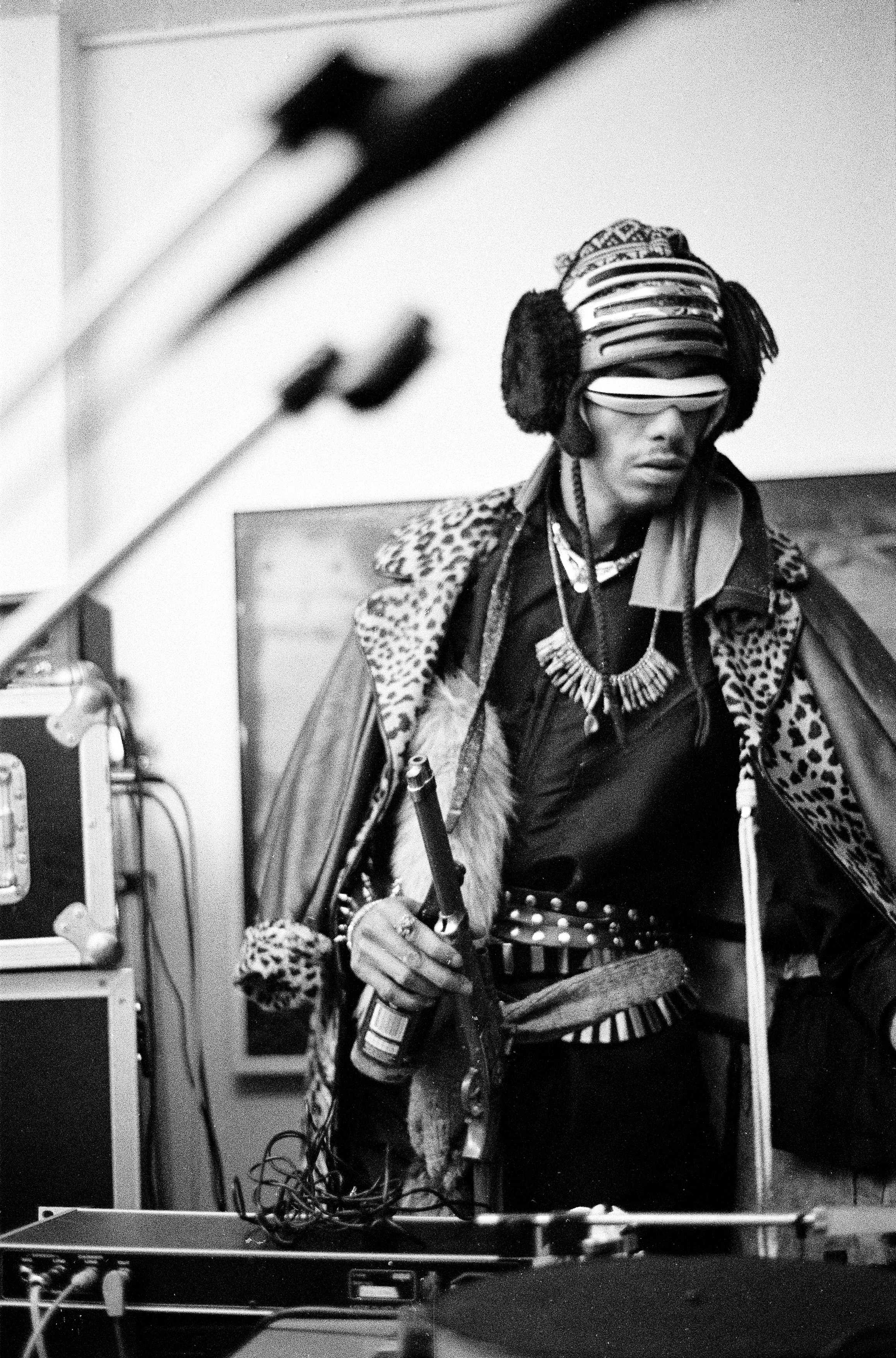 A black and white photo of Rammellzee preforming while wearing multiple headphones