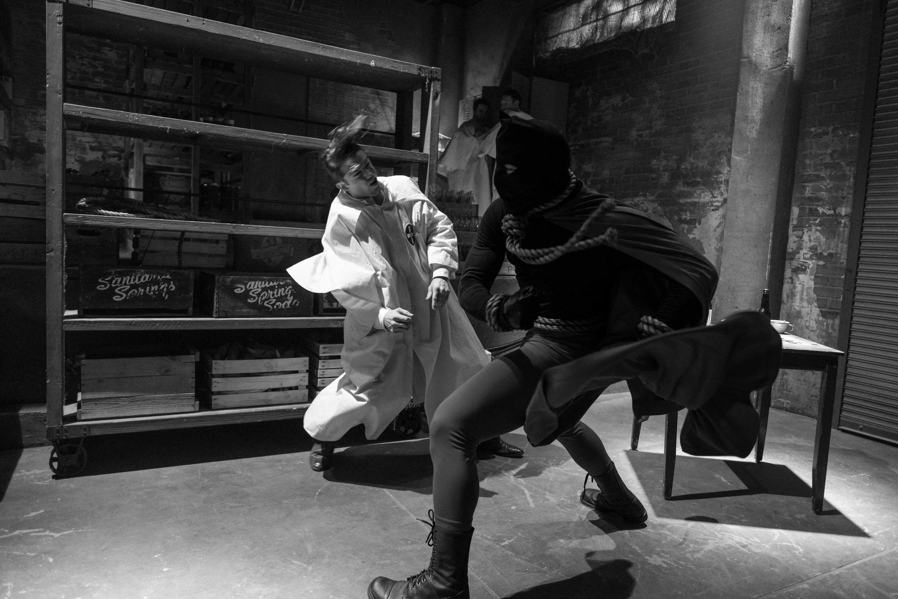 A black and white image of Hooded Justice mid fight. They are fighting a person dressed in white in a dark storage room.