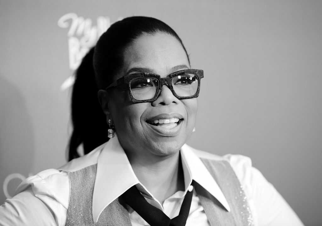 Black-and-white close-up portrait of a person with hair pulled back into a high ponytail, wearing large rectangular eyeglasses, a collared shirt, tie, and vest.