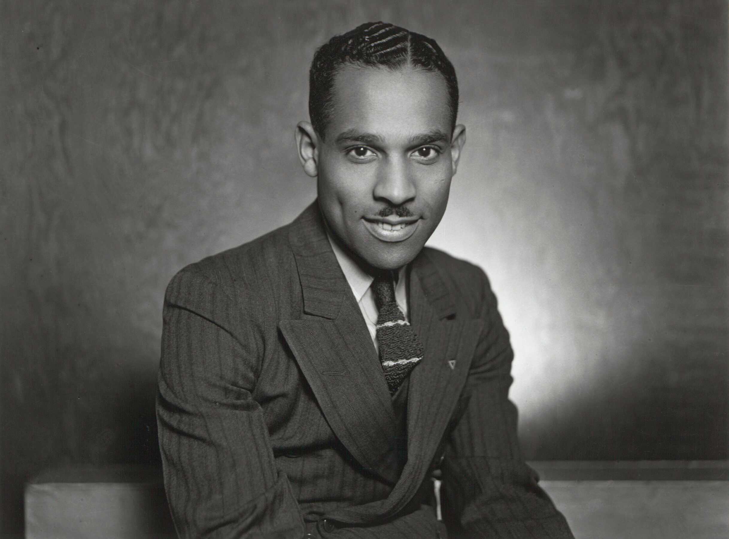 A black and white photograph of Charles "Teenie" Harris. This self-portrait pictures Harris from the waist up, seated in his photography studio, wearing a striped suit.