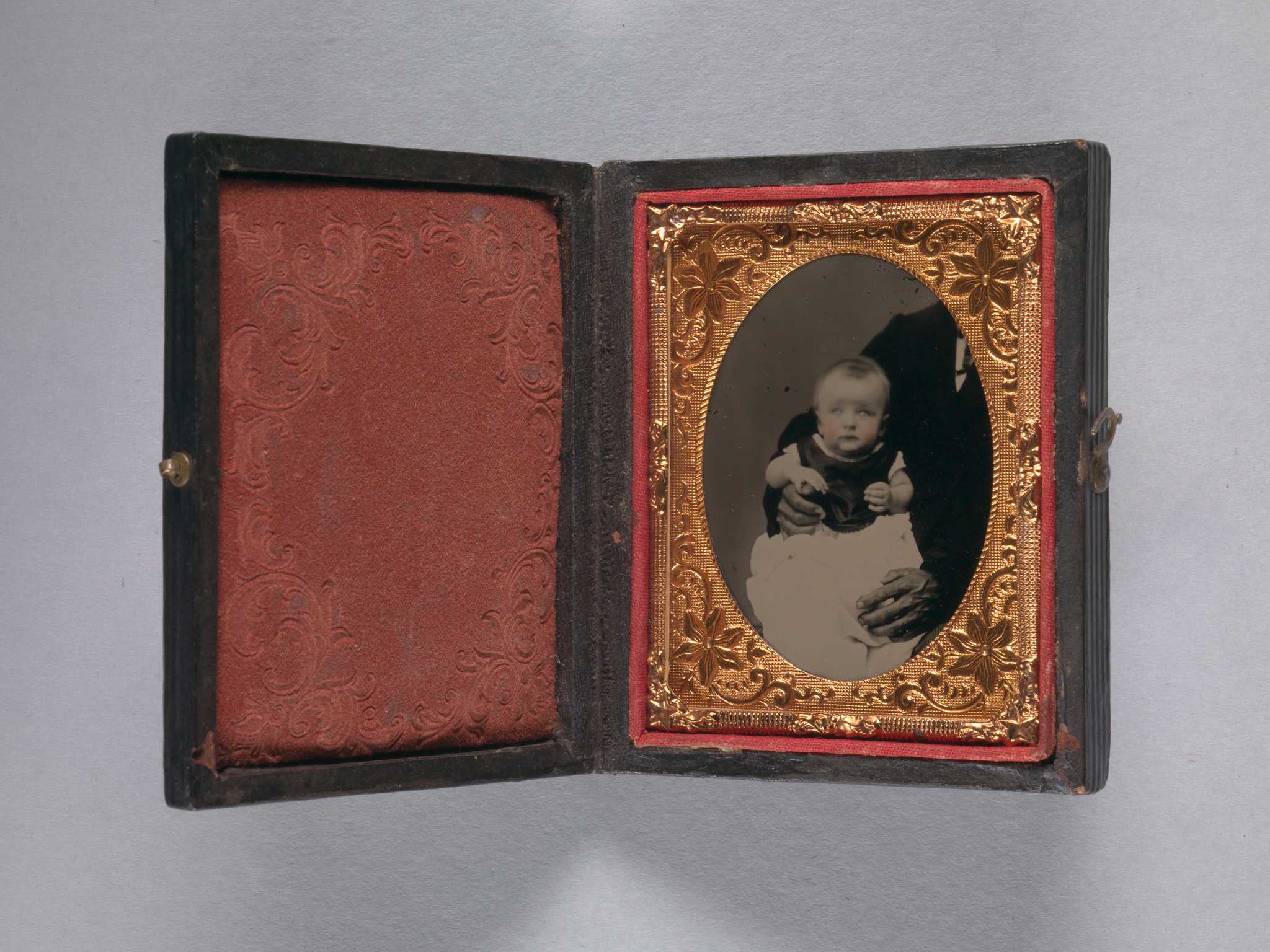 This 1/9 plate cased ambrotype depicts a white baby being held for the camera by a black man whose shoulder and hands are visible, but is otherwise unseen. The baby's cheeks have been tinted pink. The photograph is in a decorative copper-colored metal oval frame with a floral, star, and scroll motif and is held in a brown case with red textile lining. There are decorative impressions on the front and back of the case, which fastens with a small metal hook and eye closure along one edge.