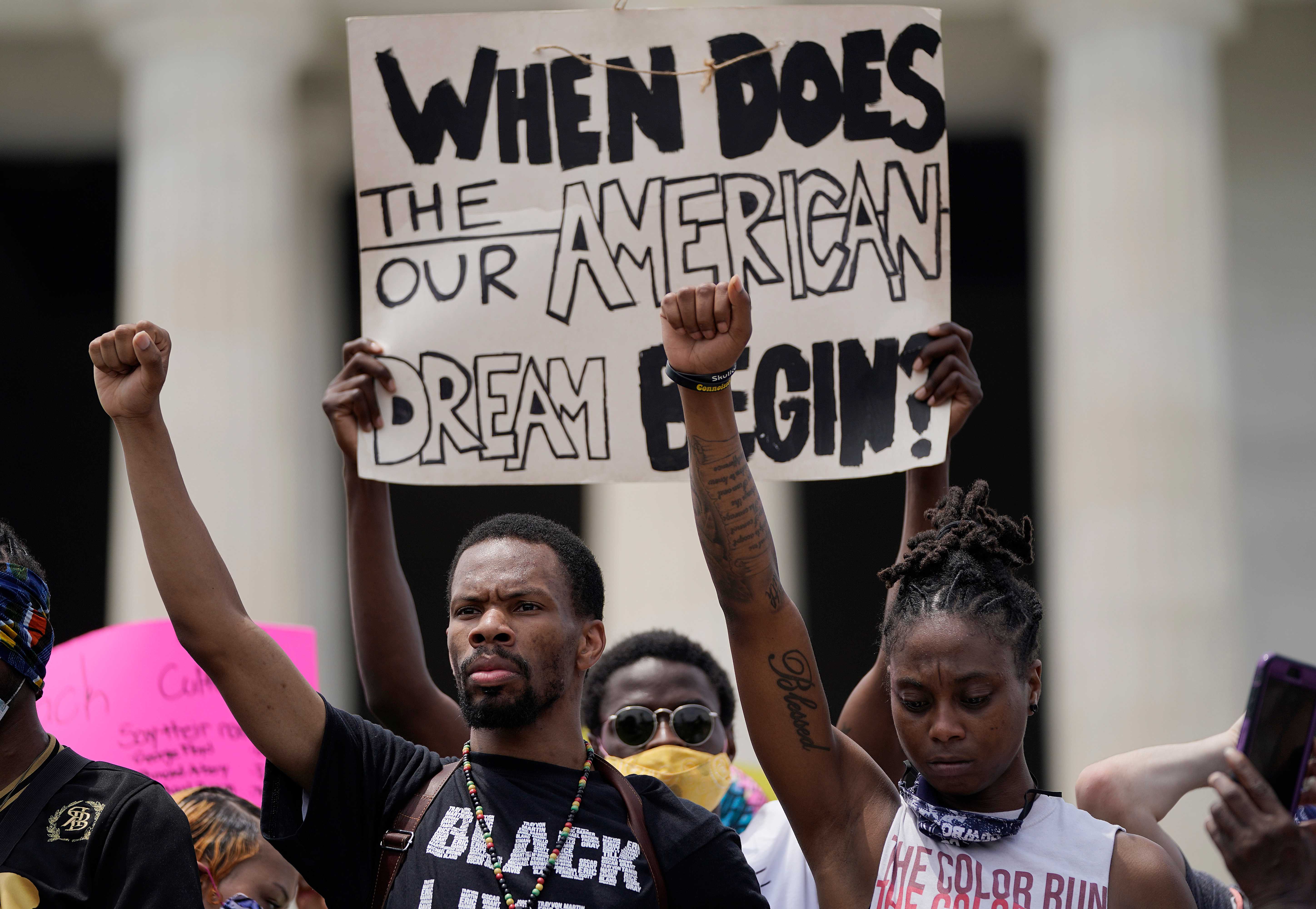 Color photograph of protesters with fists raised with sign reading "When Does The/Our American Dream Begin?"
