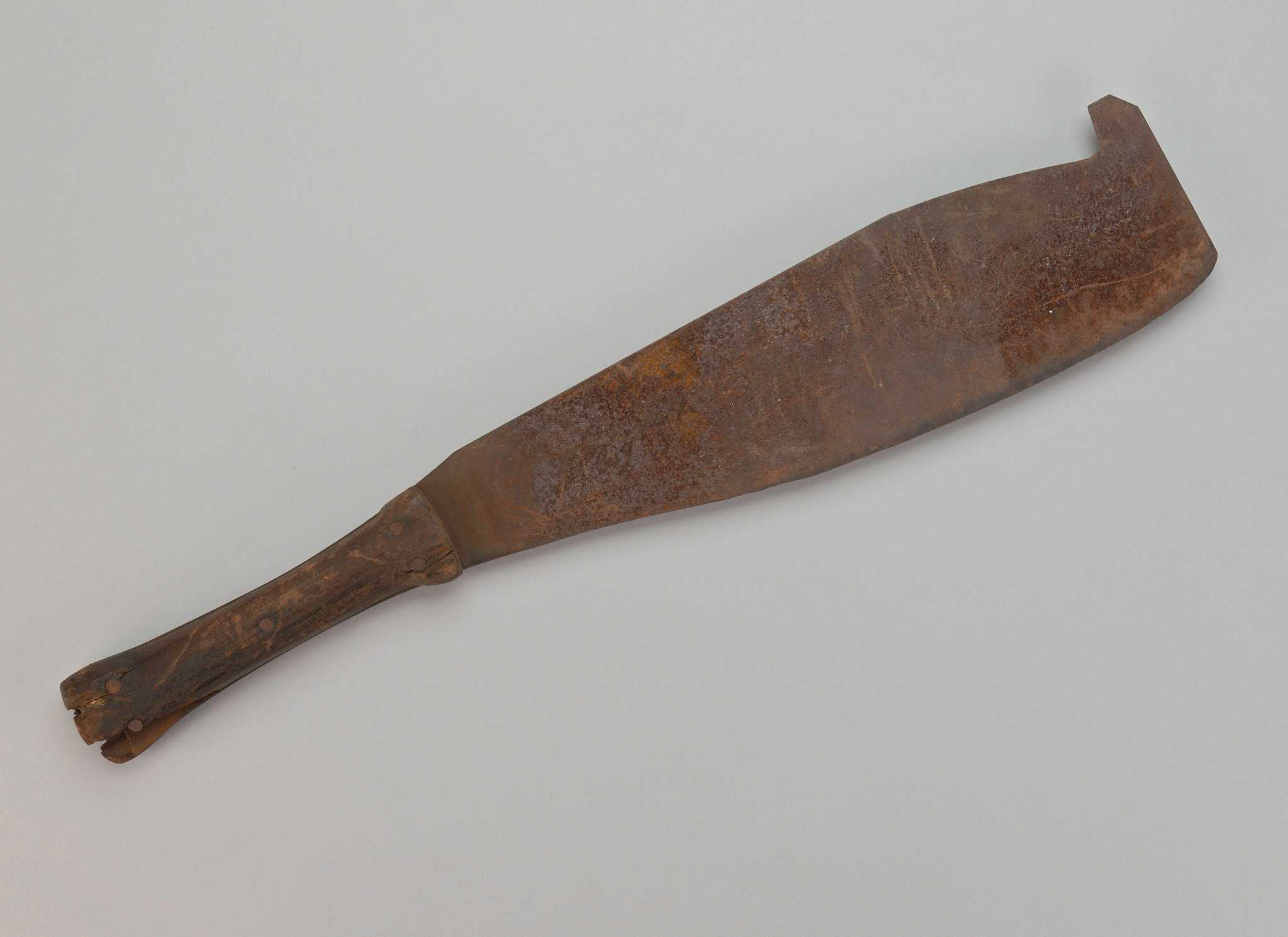 A short-handled sugar cane cutter with metal blade and wood handle. The metal blade is flat and wide at the head, tapering slightly towards the hilt. There is a hook protruding from the widest portion of the head of the blade. There are four metal nails in the wood handle. There are fragments of wood missing from the butt of the handle, exposing parts of the nails.