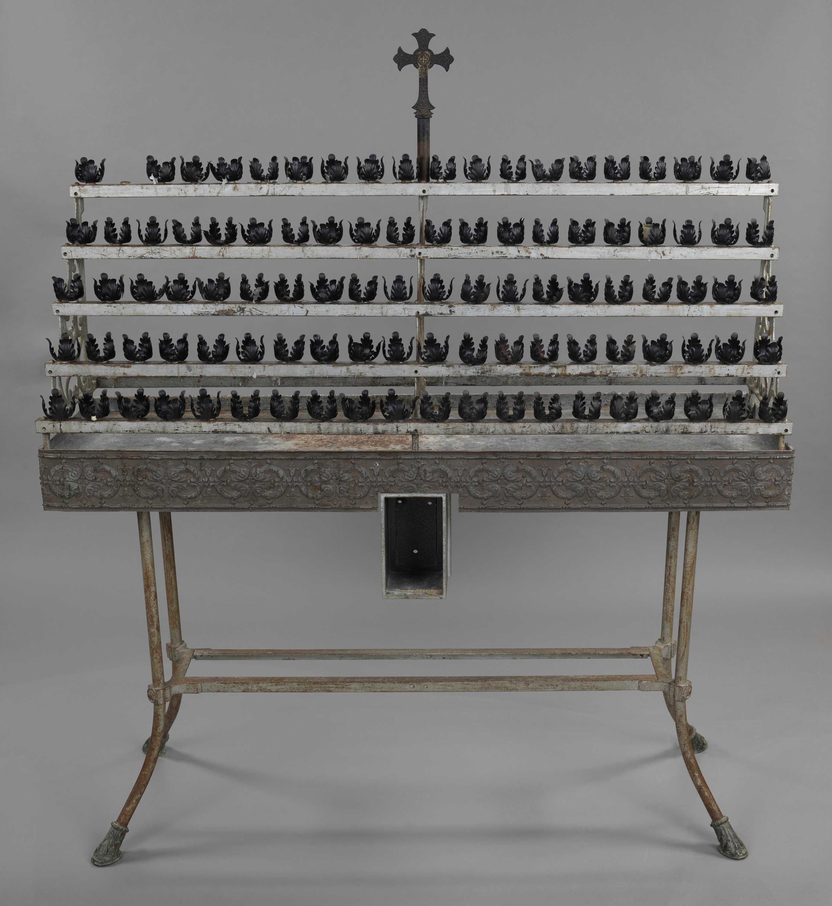 A votive candle stand with ninety-nine candle holders. The holders all feature four stylized leaves. The top of the stand has five rows of with twenty holders each. One of the holders on the top row is missing. The stand is supported by an inverted arch made up of scrollwork with stylized vines on the outside edges and center. The supports are attached to rows of votive holders by screws. The stand is topped with cross. The cross sits on top of a plain column. The cross is capped at the top, bottom, and either arm with trefoils. At the intersection of the arms and panel are two concentric circles with the outside circle appearing behind the cross and the inner circle inside the intersection. Within the inner circle of the intersection is a small Greek cross. The surface of the cross is covered with stylized foliage and leaves. 

The metal base has a stylized floral frieze running around the entire face. Within each set of stylized leaves is a Greek cross. The top and bottom of the frieze has a dashed line separated by two dots. The stand top has a small lipped edge where the votive holder rack sits. The base is supported by four curved metal legs. The legs curve away from the base and are capped with stylized foliage for feet. The legs are supported with rectangular bars midway down the legs. A floral decoration is found where the bar supports attach to the legs, with centers that are screws.


A 10mm Craftsman double-headed wrench was found inside the candle stand during conservation treatment.